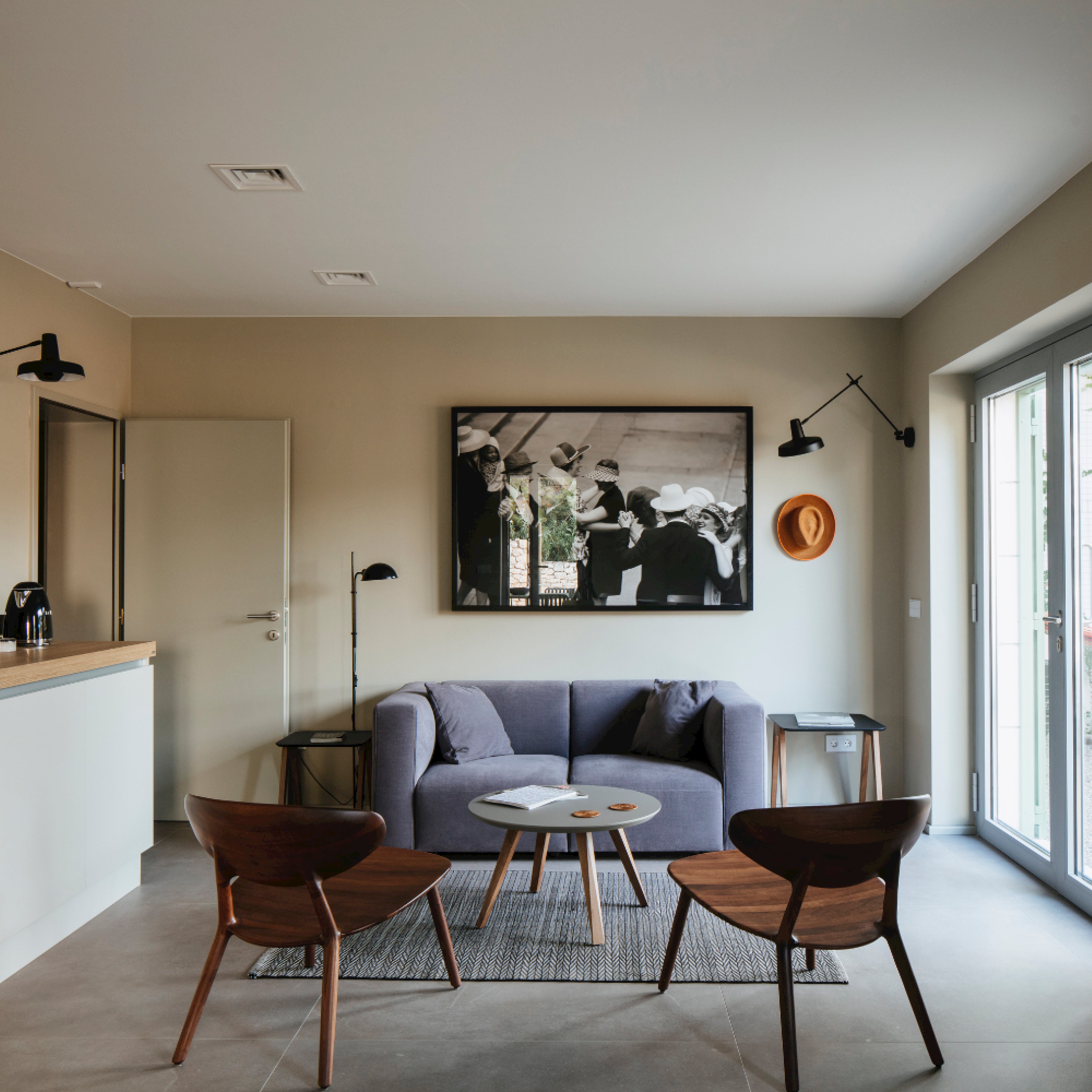 A modern living room showcases a gray sofa, two Artisan WU Lounge Chairs made from solid wood, and a small round table on a rug. A black-and-white photo hangs above the sofa. Wall lamps adorn the light-colored walls, where a straw hat also hangs. Large windows illuminate the luxury wood texture of the décor.