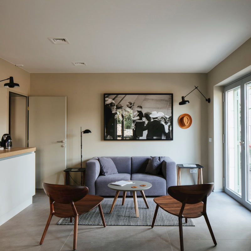 A modern living room showcases a gray sofa, two Artisan WU Lounge Chairs made from solid wood, and a small round table on a rug. A black-and-white photo hangs above the sofa. Wall lamps adorn the light-colored walls, where a straw hat also hangs. Large windows illuminate the luxury wood texture of the décor.