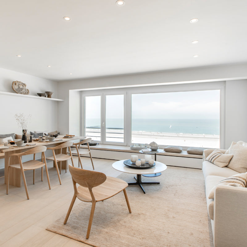 A modern living room with ocean views features a beige sofa, Artisans WU Lounge Chairs crafted from light solid wood, a round coffee table, and a dining table. Minimal decor in neutral hues enhances the serene seaside ambiance.