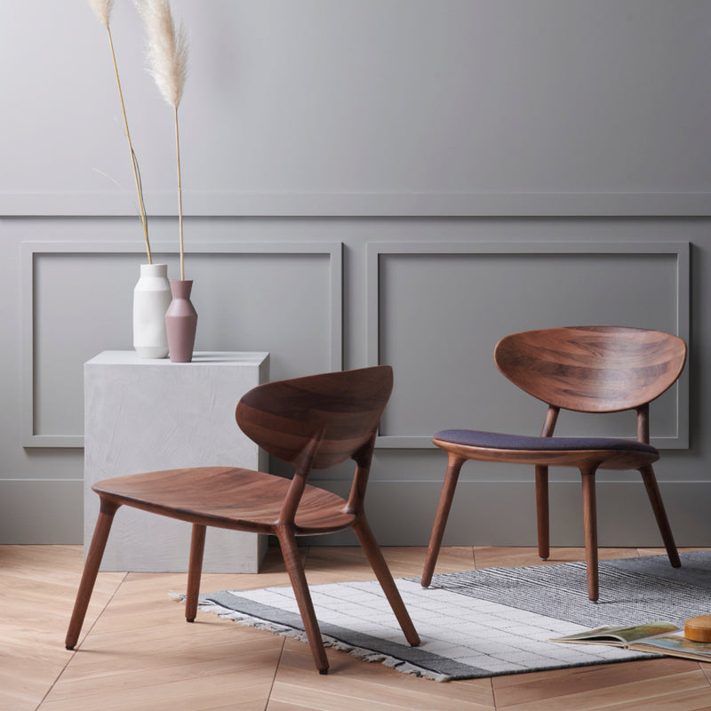 Two WU Lounge Chairs by Artisan, crafted from luxury wood textures, adorn a herringbone-patterned floor beside a textured white pedestal holding vases and tall pampas grass. The elegant, minimalist setting features a neutral palette with wall molding, while an open book rests on a nearby rug.