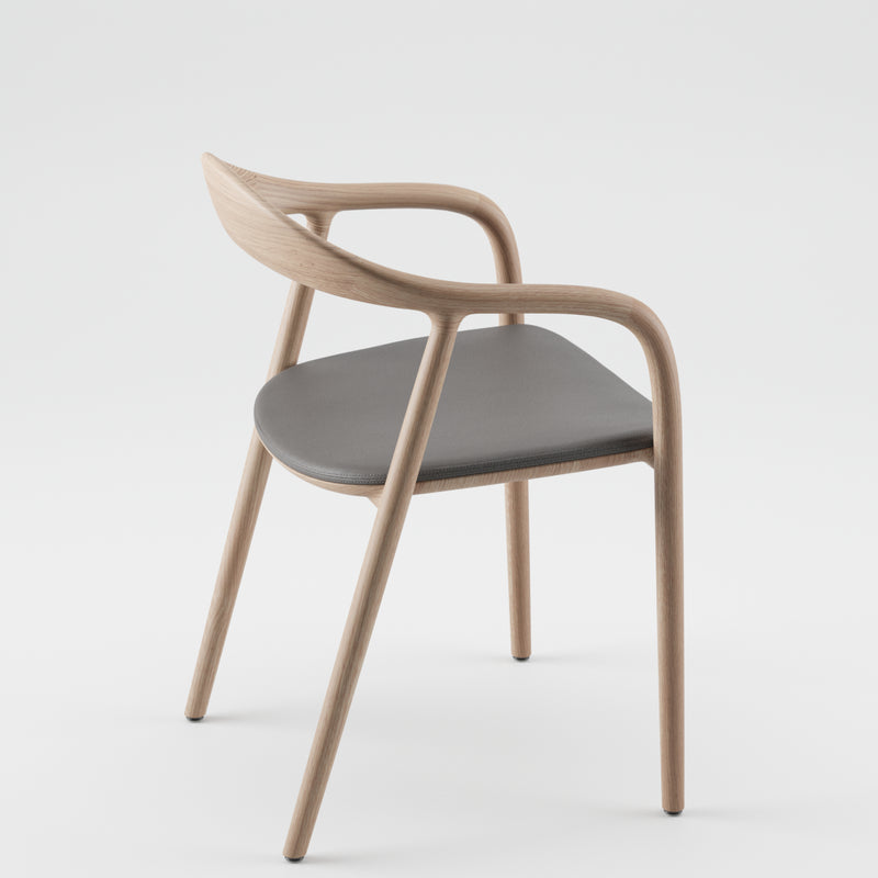 Artisans NEVA chair boasts a sleek design with a smooth American walnut frame crafted using 5-axis CNC technology, featuring gently curved armrests and legs. It includes a dark cushioned seat and is set against a light background for contrast.