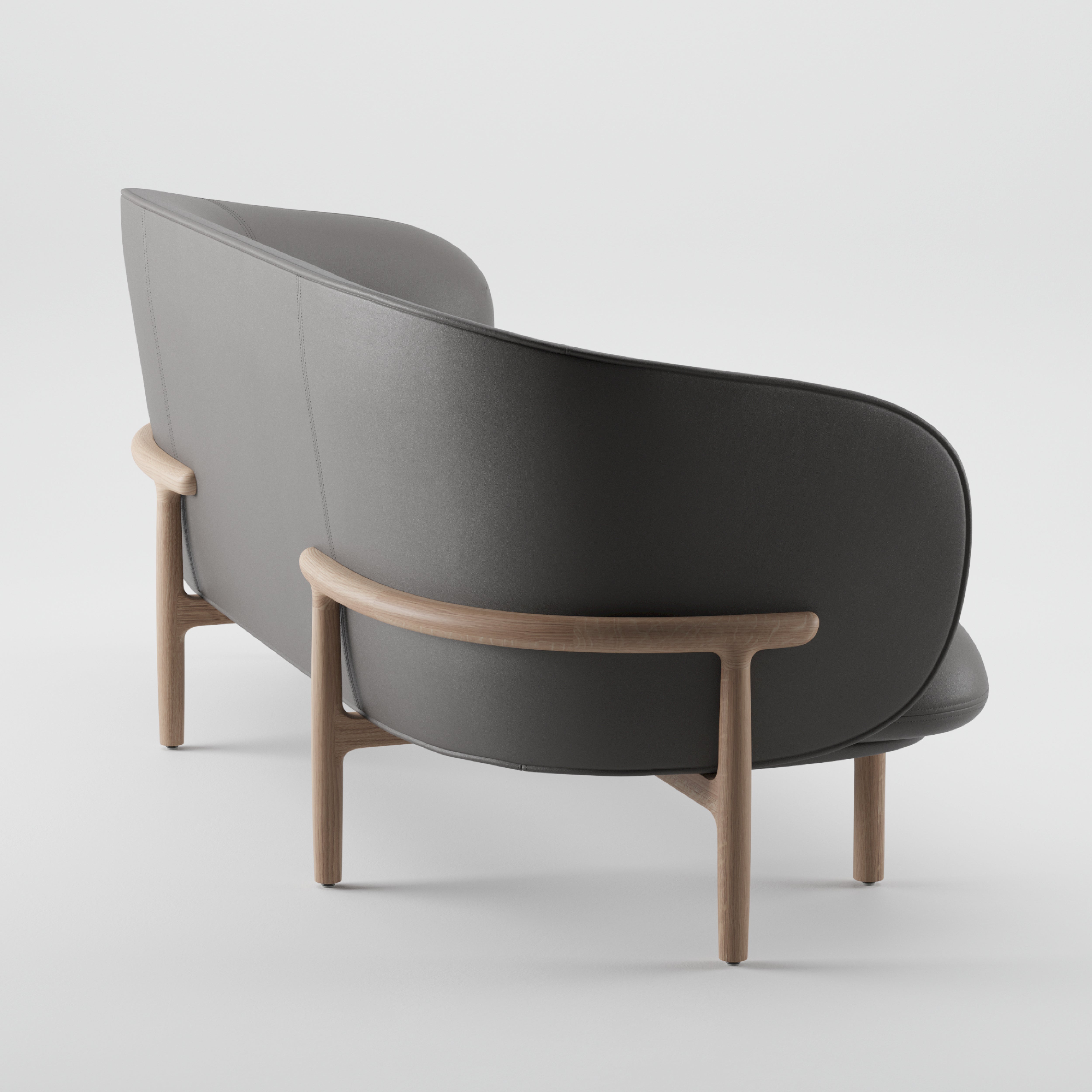 Discover the MELA Two Seater by Artisan, a modern minimalist sofa with a smooth grey upholstered shell and curved design. Its light wooden frame and legs provide mid-century elegance, creating a sleek look against a plain white backdrop.