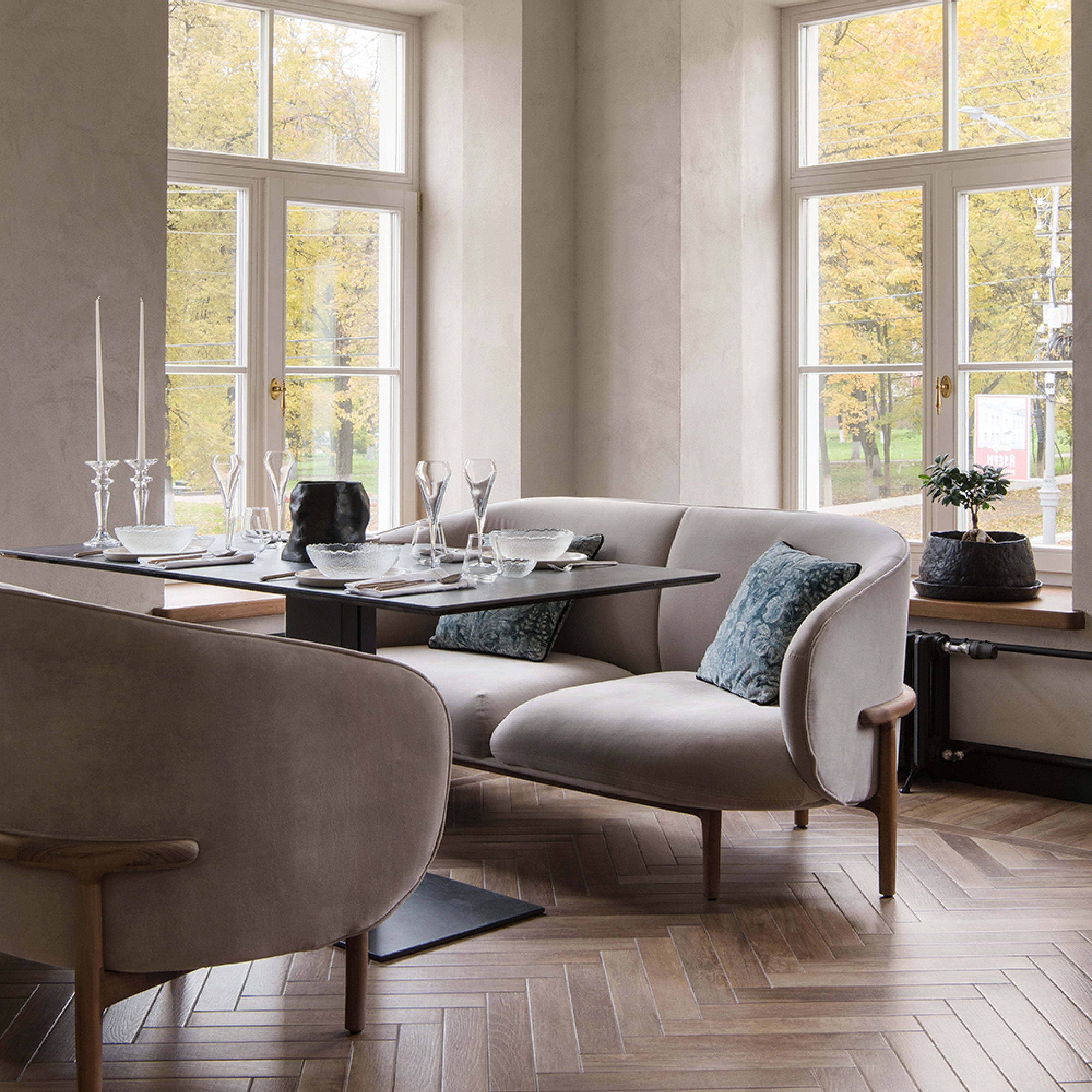 A cozy dining area features Artisans light gray upholstered MELA Two Seater around a dark table with elegant tableware and crystal candlesticks. Large windows offer views of autumn trees, while a small potted plant sits on the window bench. A herringbone-patterned floor enhances the spaces charm.