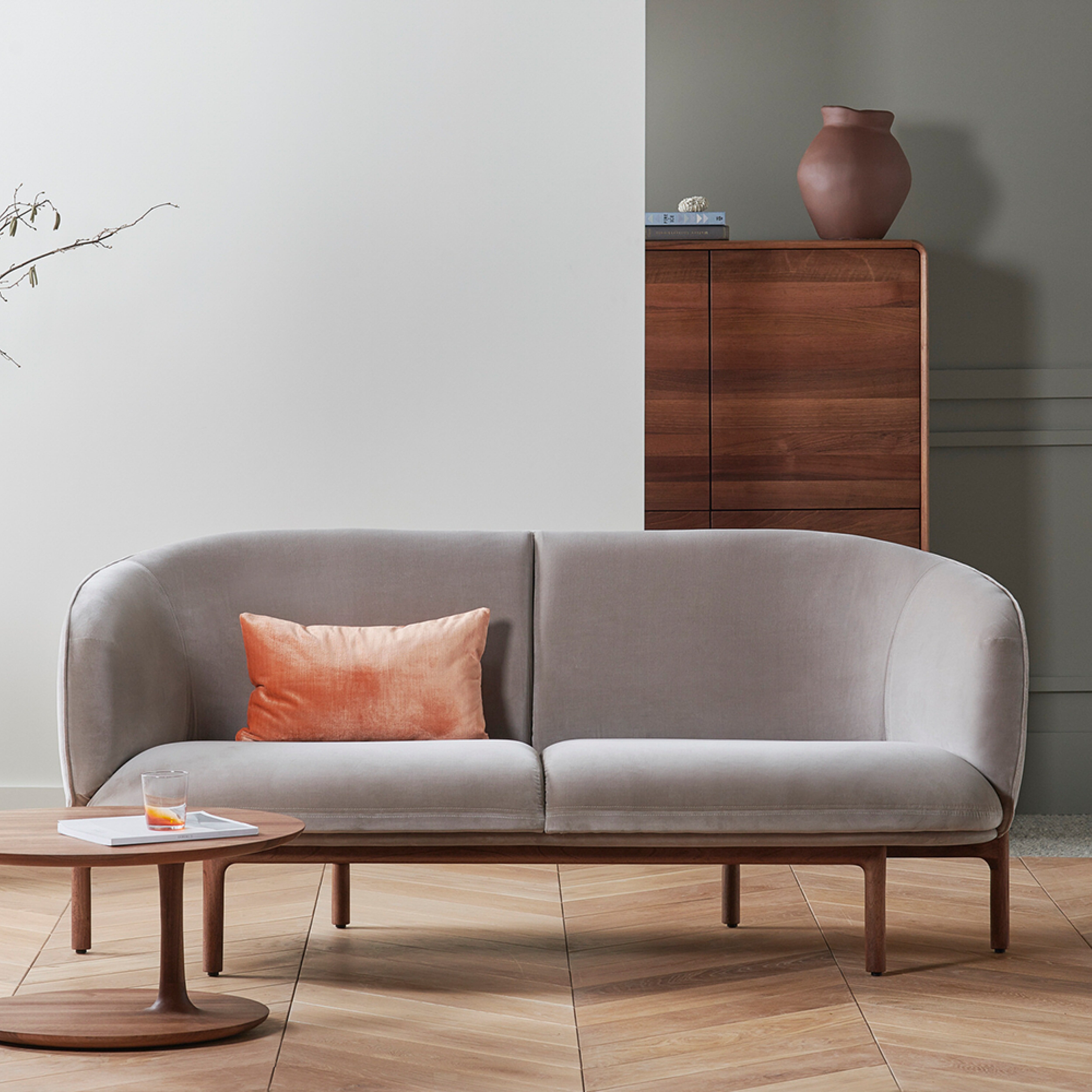 A modern living room showcases the light gray MELA Two Seater by Artisan with a peach pillow. A round wooden coffee table has a glass and magazine, while a wooden cabinet with books and a vase stands in the background, set against two-tone walls and herringbone wood flooring.