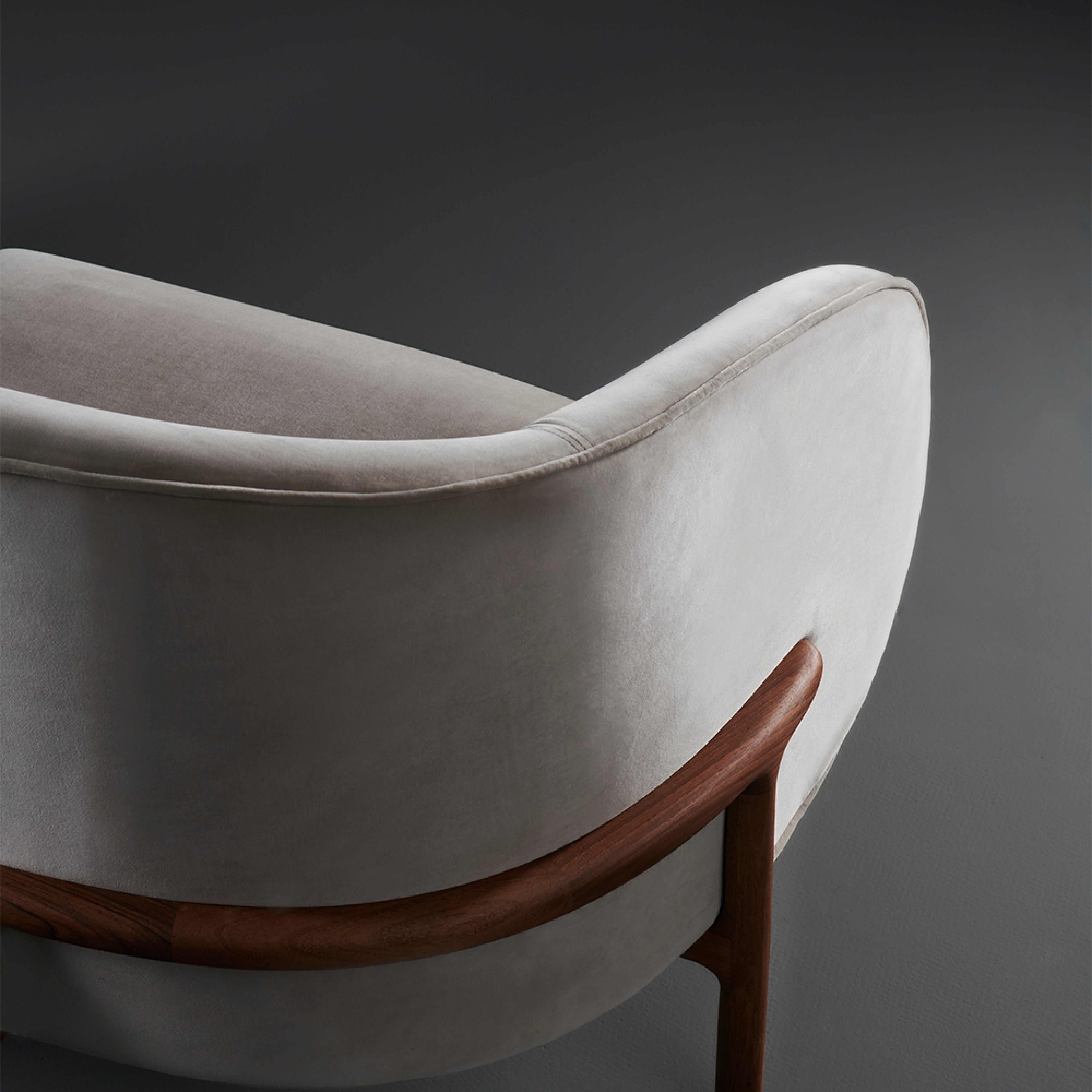 Elegant and modern, the MELA Two Seater by Artisan features a smooth, light grey upholstered backrest and seat. Its curved wooden frame has a rich brown finish, offering a striking contrast against dark settings.
