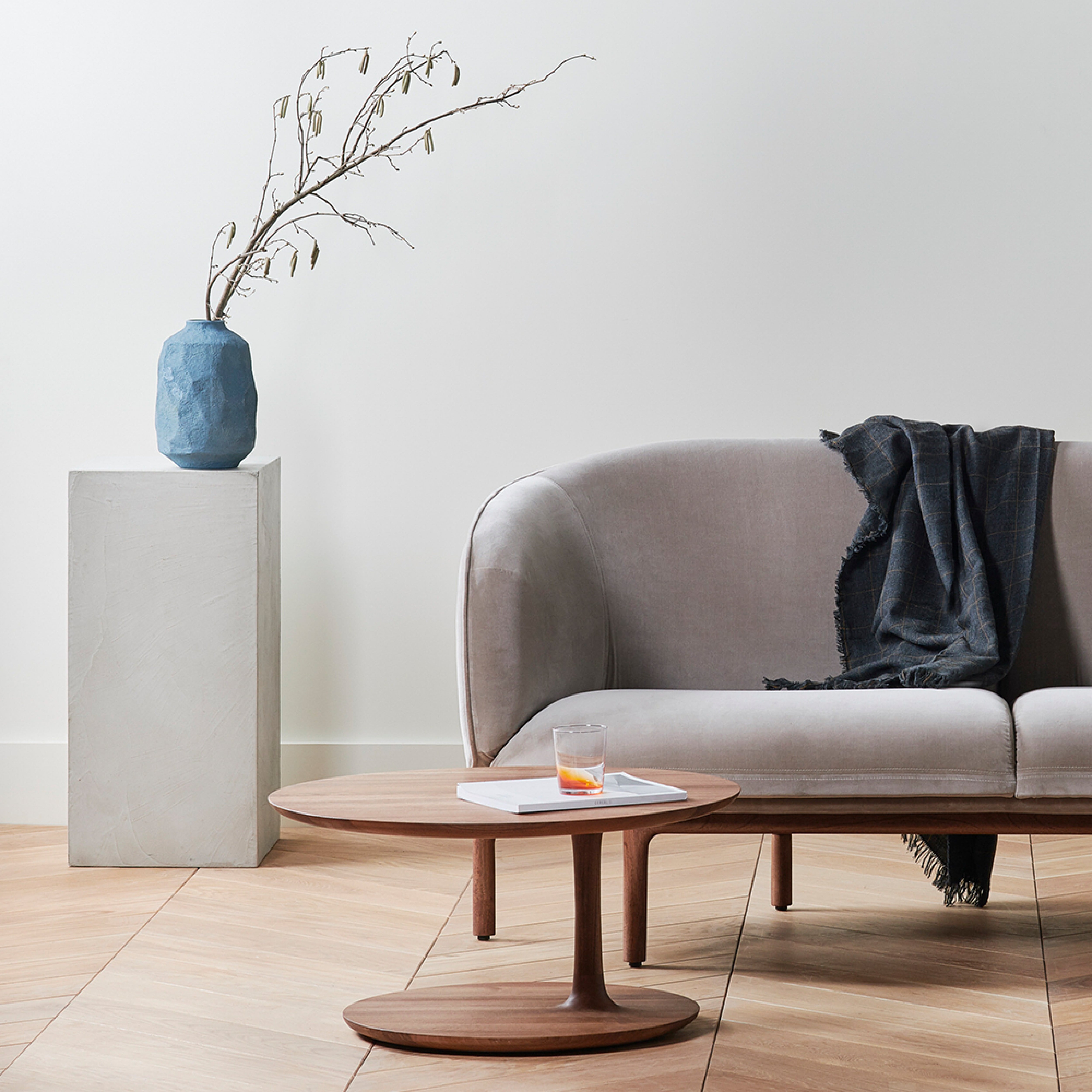 A modern living room features an Artisan MELA Two Seater in gray and an upholstered chair. A wooden coffee table holds a magazine and glass of water, while a tall pedestal displays a blue vase with branches. A dark throw blanket draped over the sofa arm complements the light wood flooring.