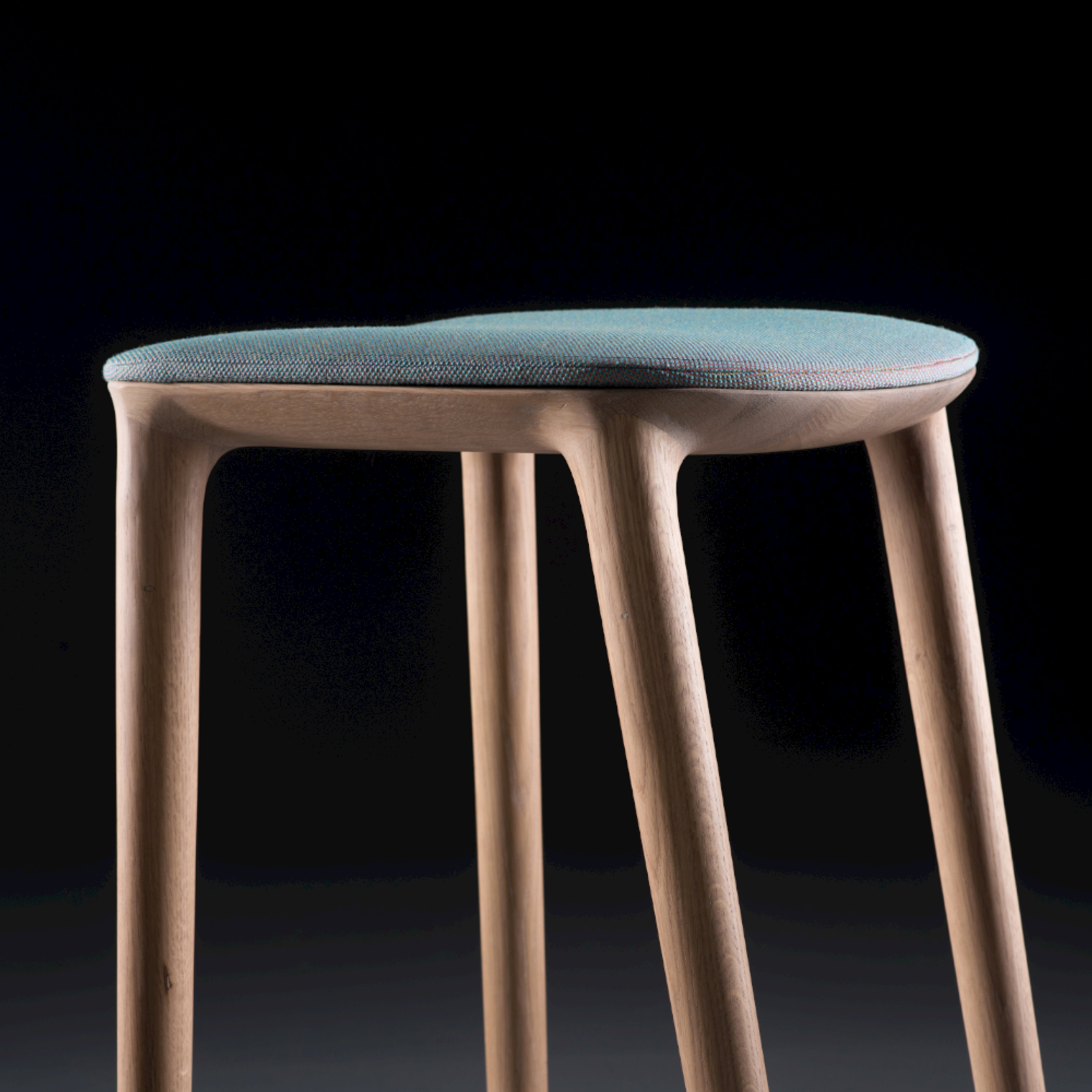 A modern NEVA barstool from Artisan, showcasing a blue textured fabric seat, sleek tapered legs, and minimalist design, highlights artisanal craftsmanship against a dark backdrop.