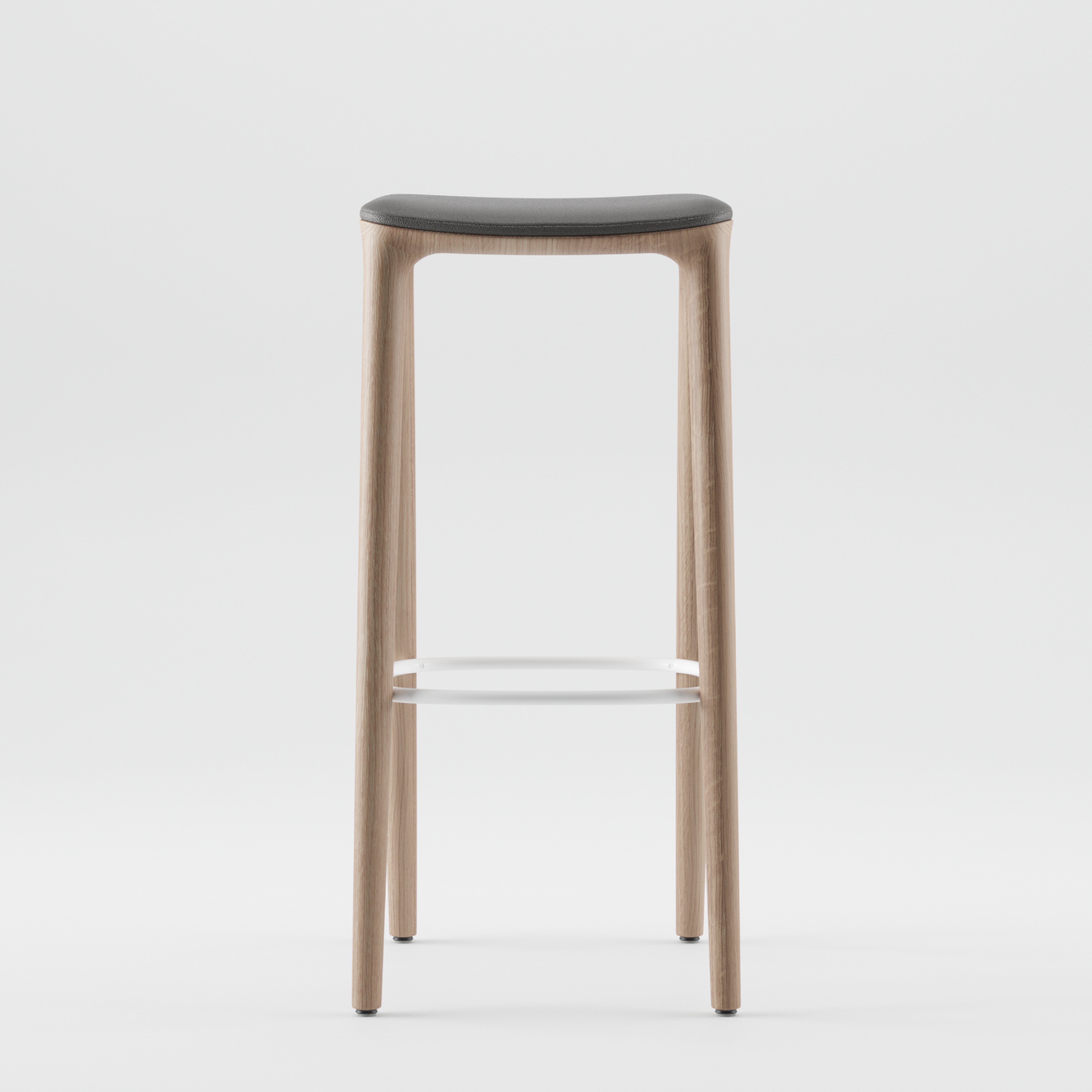 The NEVA barstool by Artisan features a minimalist design with a dark cushioned seat, four slender wooden legs, and a horizontal metal bar for stability, highlighting artisanal craftsmanship. It is showcased against a plain white background.