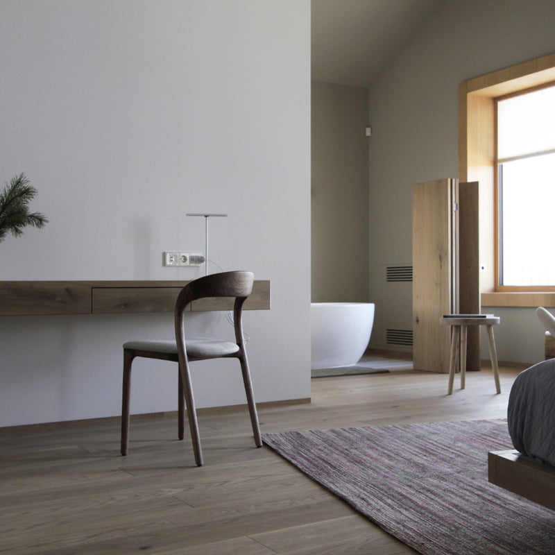 This minimalist bedroom boasts light wood flooring, a wooden desk with the Artisan NEVA light chair, and a white freestanding bathtub near a large window. It features a simple bed, textured rug, and artisanal carpentry for added elegance.