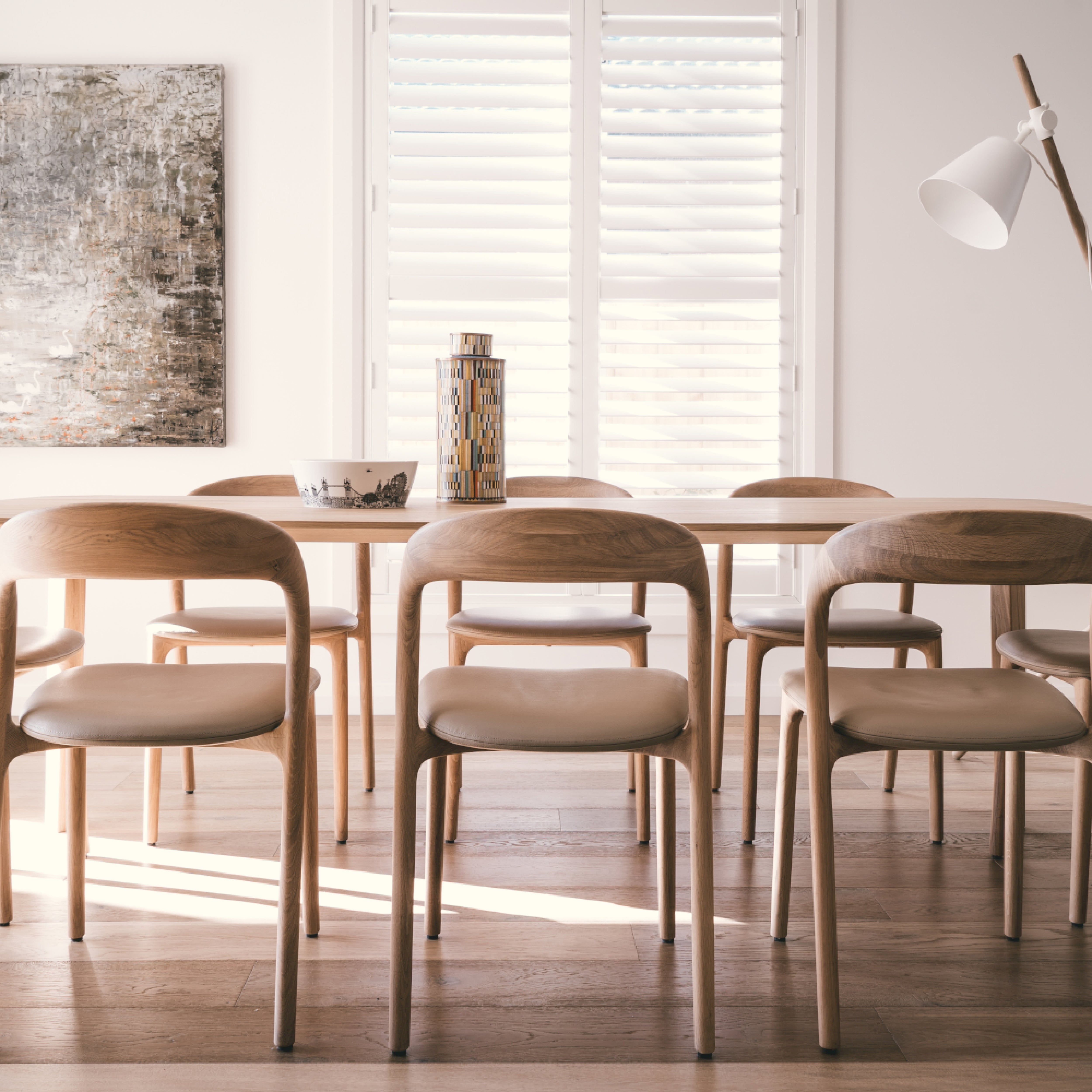 A minimalist dining room showcases a wooden table from the Neva collection, surrounded by six Artisan NEVA light chairs with cushions. A textured abstract painting adorns the wall alongside a tall lamp. Sunlight streams through white shutters, highlighting the artisanal carpentry.