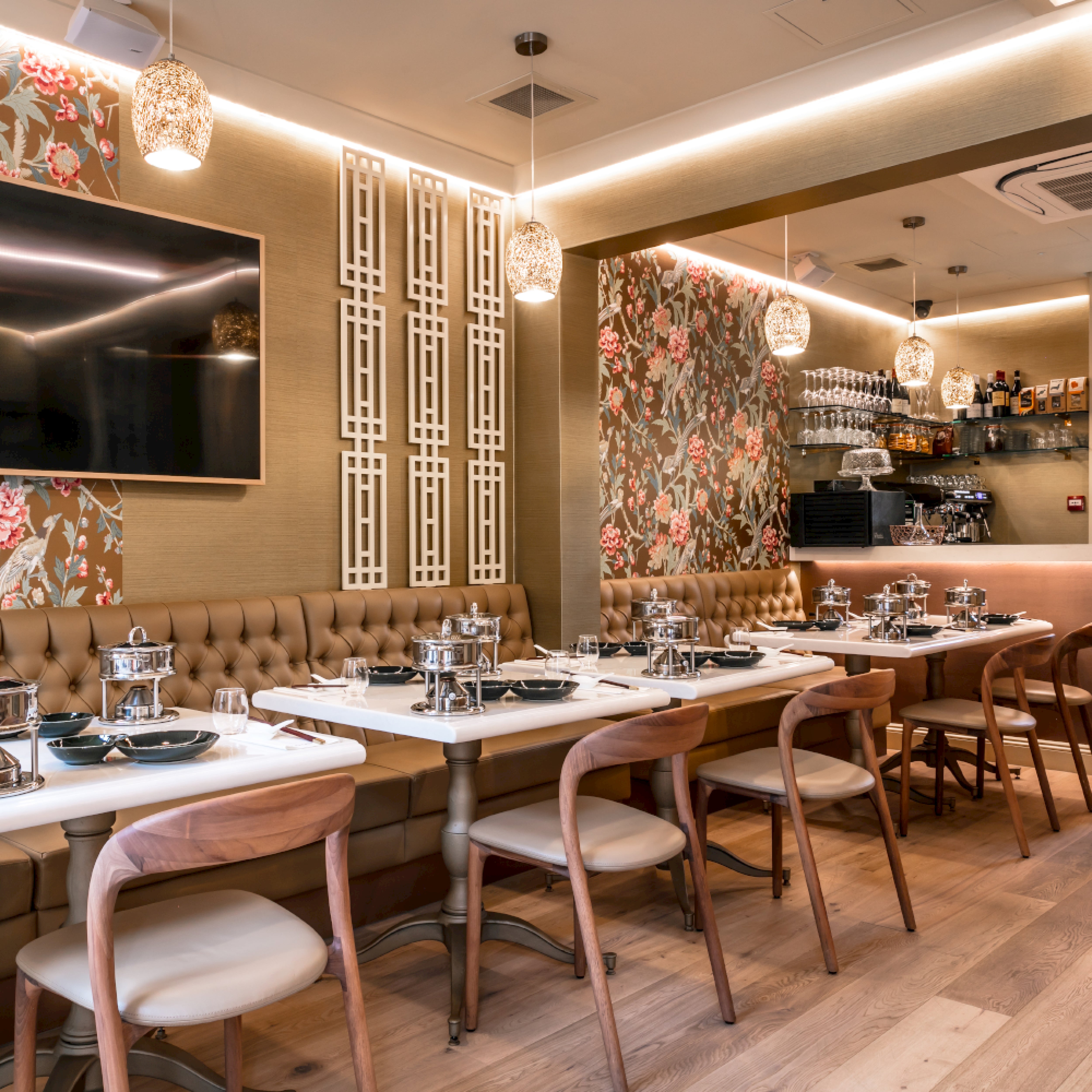 The elegant restaurant interior features floral wallpaper, a long table with tan upholstered bench seating, and Artisan-crafted NEVA light wooden chairs. Place settings include black bowls and silver hot pots. Warm lighting from pendant lamps enhances the cozy atmosphere.