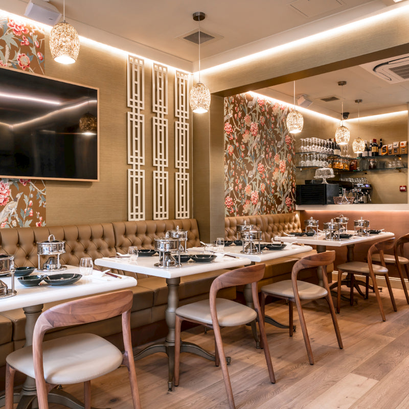 The elegant restaurant interior features floral wallpaper, a long table with tan upholstered bench seating, and Artisan-crafted NEVA light wooden chairs. Place settings include black bowls and silver hot pots. Warm lighting from pendant lamps enhances the cozy atmosphere.