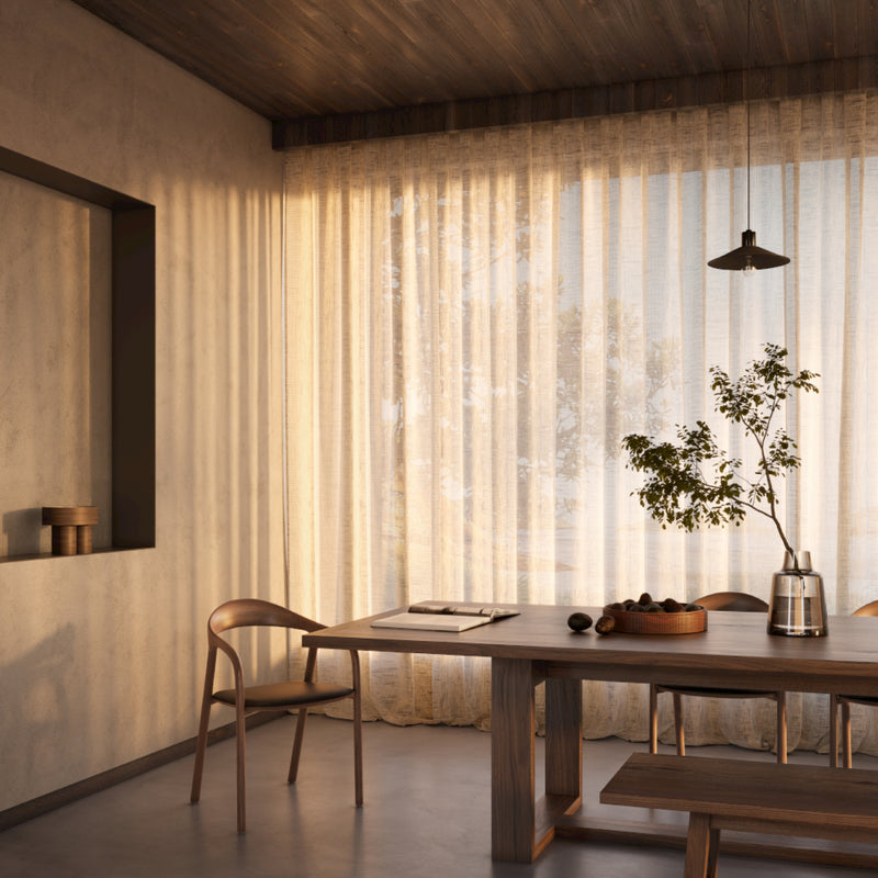 A cozy dining room features a wooden table and NEVA chairs by Artisan, crafted from rich American walnut. Soft light filters through sheer curtains, illuminating a potted plant and fruit bowl on the table. A pendant light hangs from the wooden ceiling, enhancing the warm ambiance.
