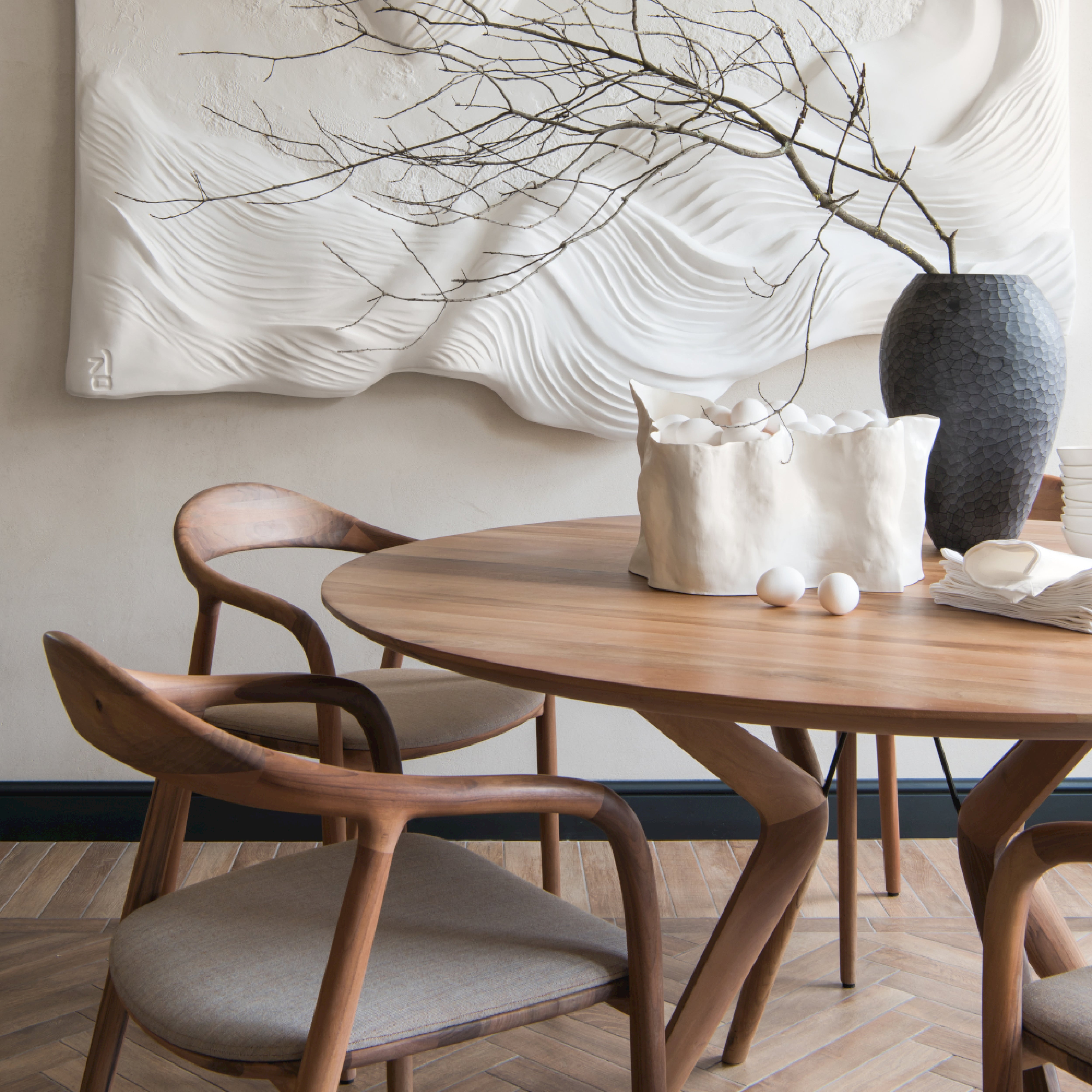 A modern dining area showcases a round wooden table with two Artisan NEVA chairs in American walnut and beige cushions. The table is adorned with a textured white ceramic pot with branches, a wrinkled white vase holding eggs, and stacked napkins. A wavy wall art piece hangs nearby.