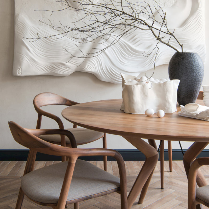 A modern dining area showcases a round wooden table with two Artisan NEVA chairs in American walnut and beige cushions. The table is adorned with a textured white ceramic pot with branches, a wrinkled white vase holding eggs, and stacked napkins. A wavy wall art piece hangs nearby.