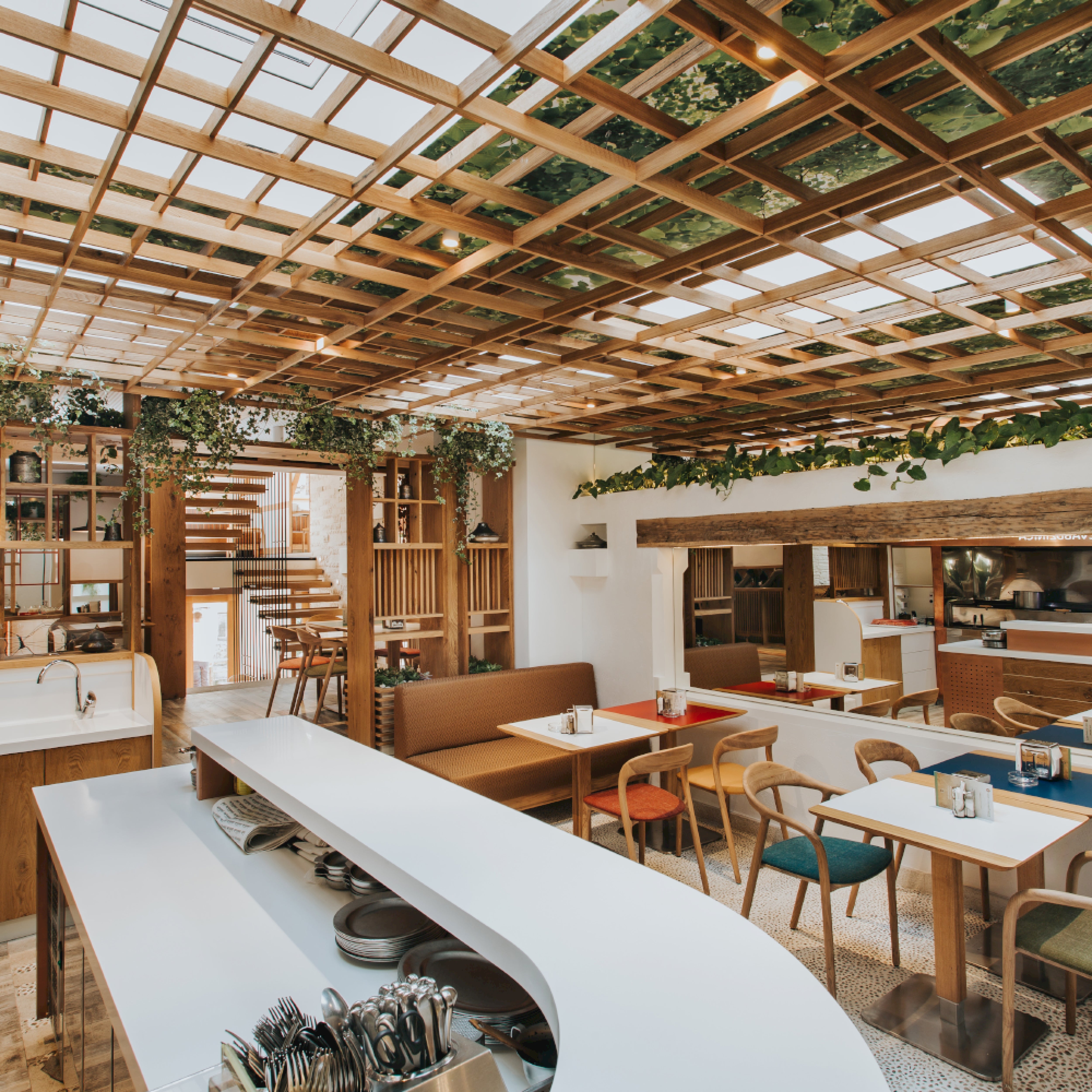 The restaurants modern interior features a wooden lattice ceiling with greenery, a long white counter, Artisan NEVA chairs, and mixed seating. An American walnut staircase adds elegance while natural light creates a welcoming atmosphere.