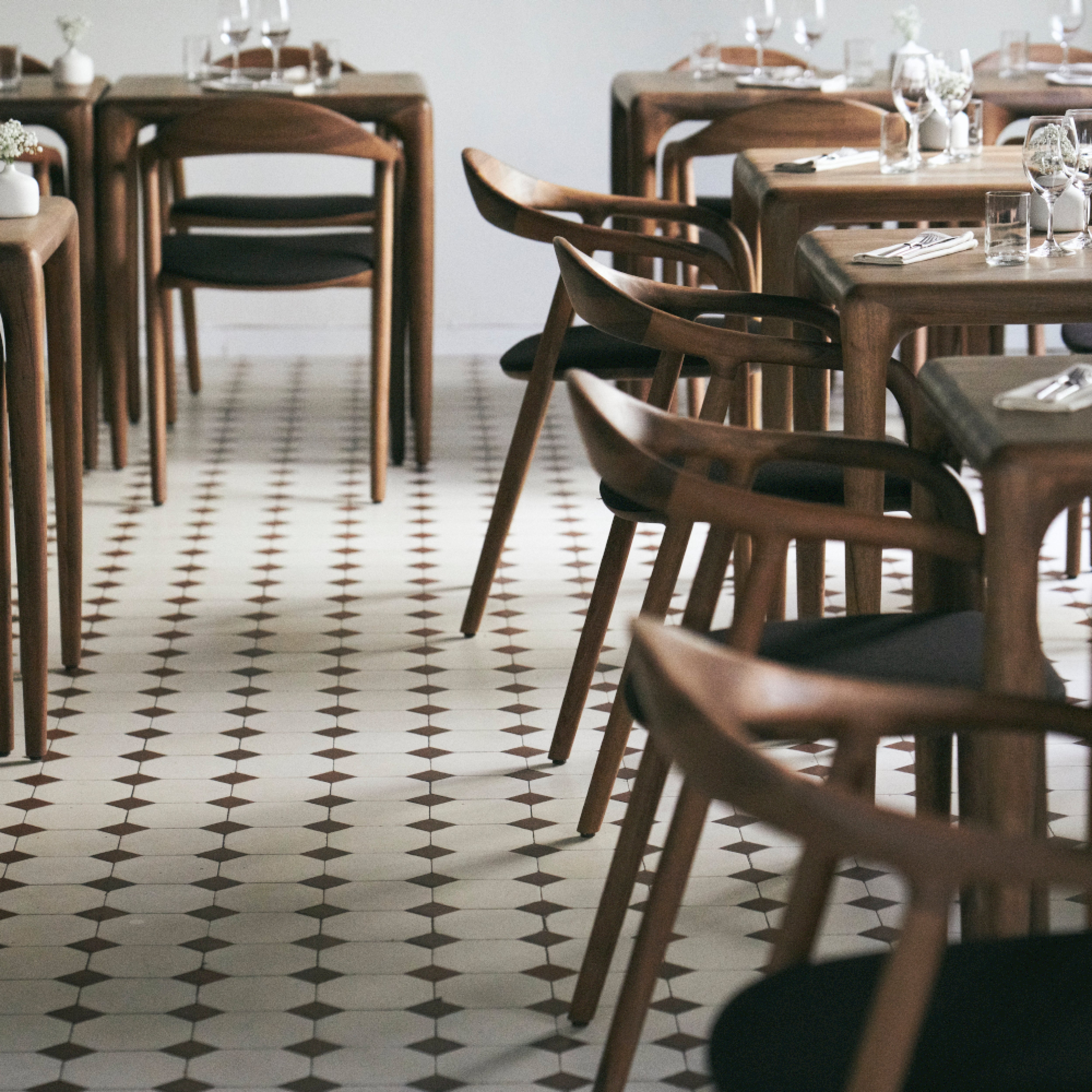 The stylish restaurant features elegant NEVA chairs by Artisan, crafted from American walnut, set around tables adorned with crisp white napkins and glassware. The geometric black and white tile flooring complements the soft lighting, enhancing the inviting atmosphere.