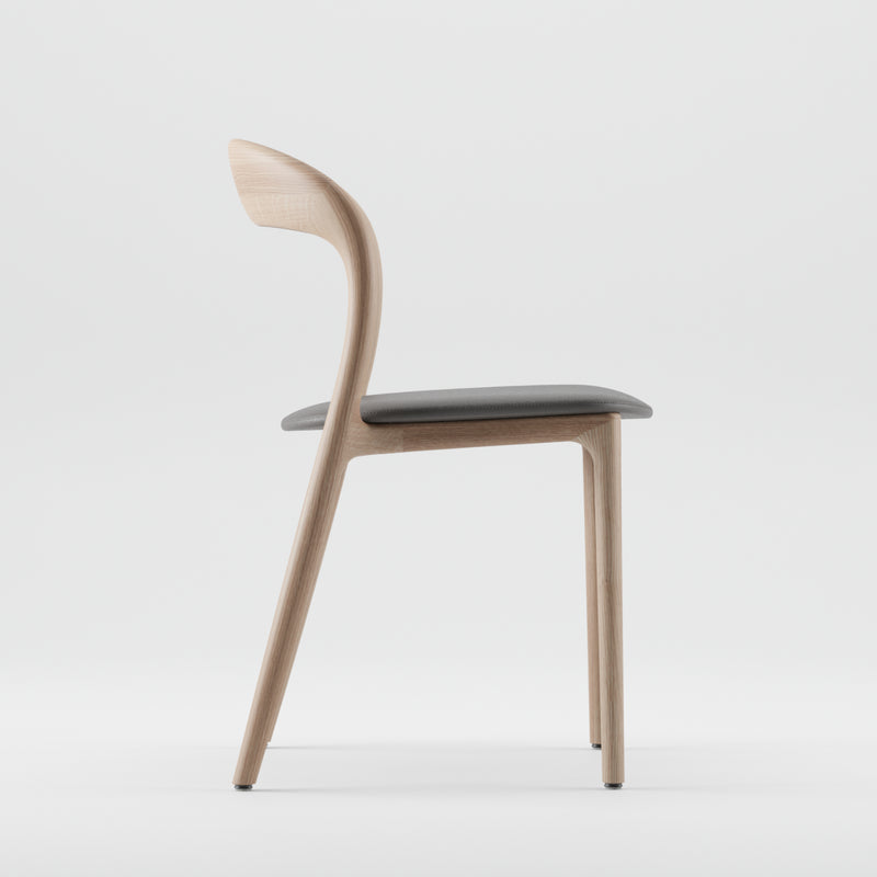 Side view of the modern NEVA light chair by Artisan, featuring a minimalist design with a curved wooden backrest, artisanal carpentry, dark cushioned seat, and sleek wooden legs against a plain white background.