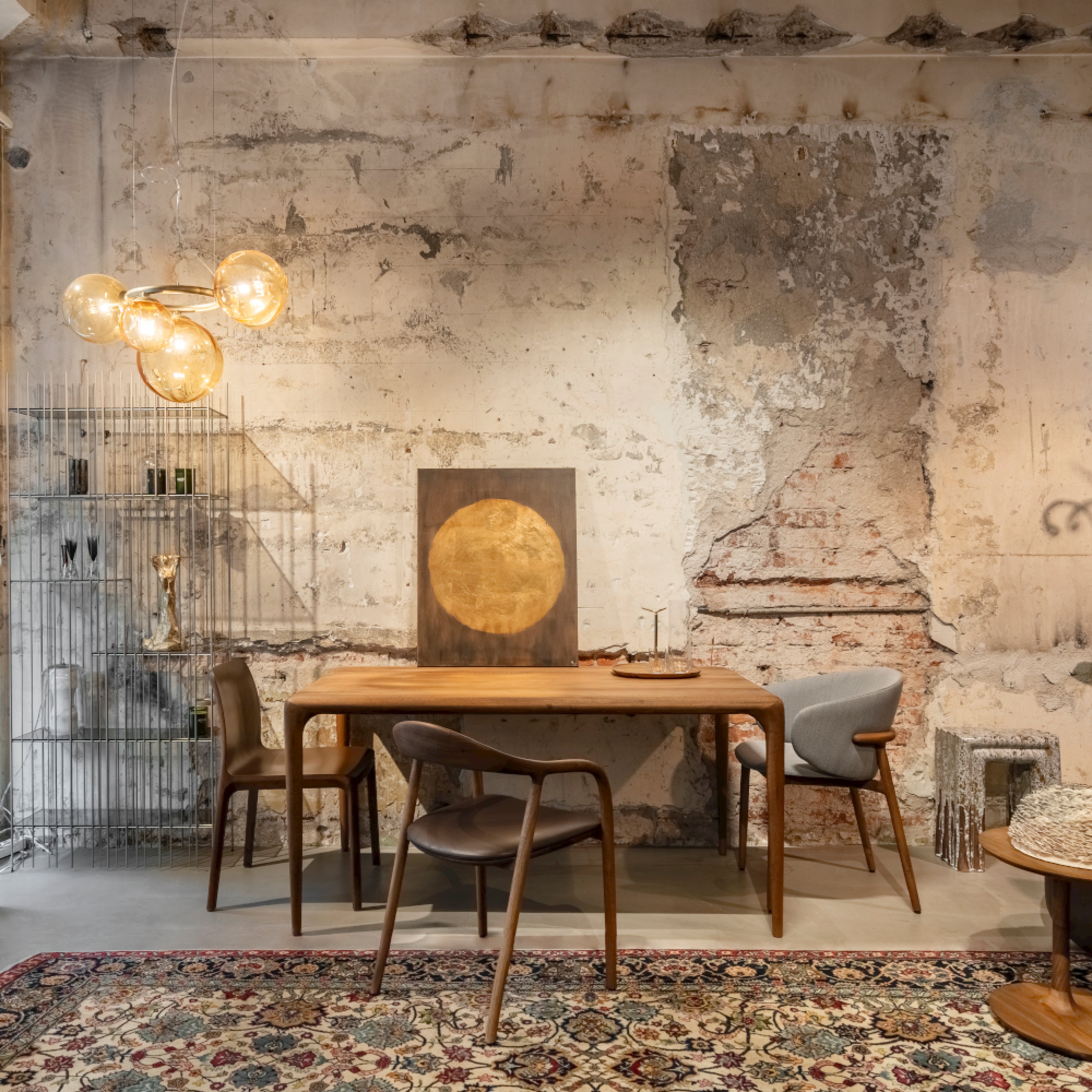 A rustic interior features a distressed wall with exposed brick, complemented by an American walnut table and Artisans NEVA chairs. A framed circular artwork adorns the wall, while a cage metal shelf and ornate rug add eclectic style. Glass orb lights overhead illuminate the space.