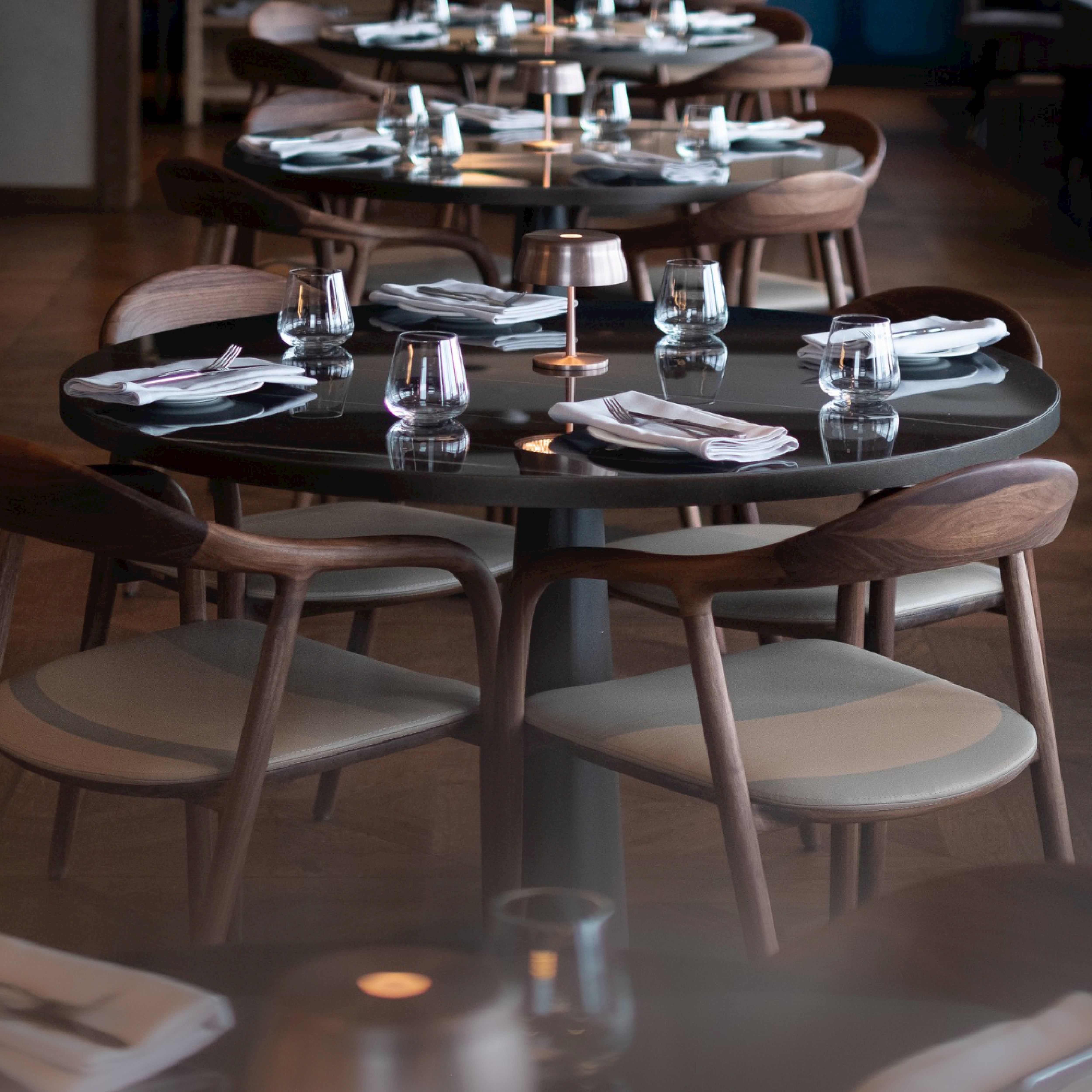 In a minimalist restaurant setting, round wooden tables feature the Artisan NEVA Easy Chair, combining ergonomic design with style. Each table is set with four water glasses, a small lamp, and neatly folded white napkins. Cushioned seats add to the warm and inviting atmosphere.