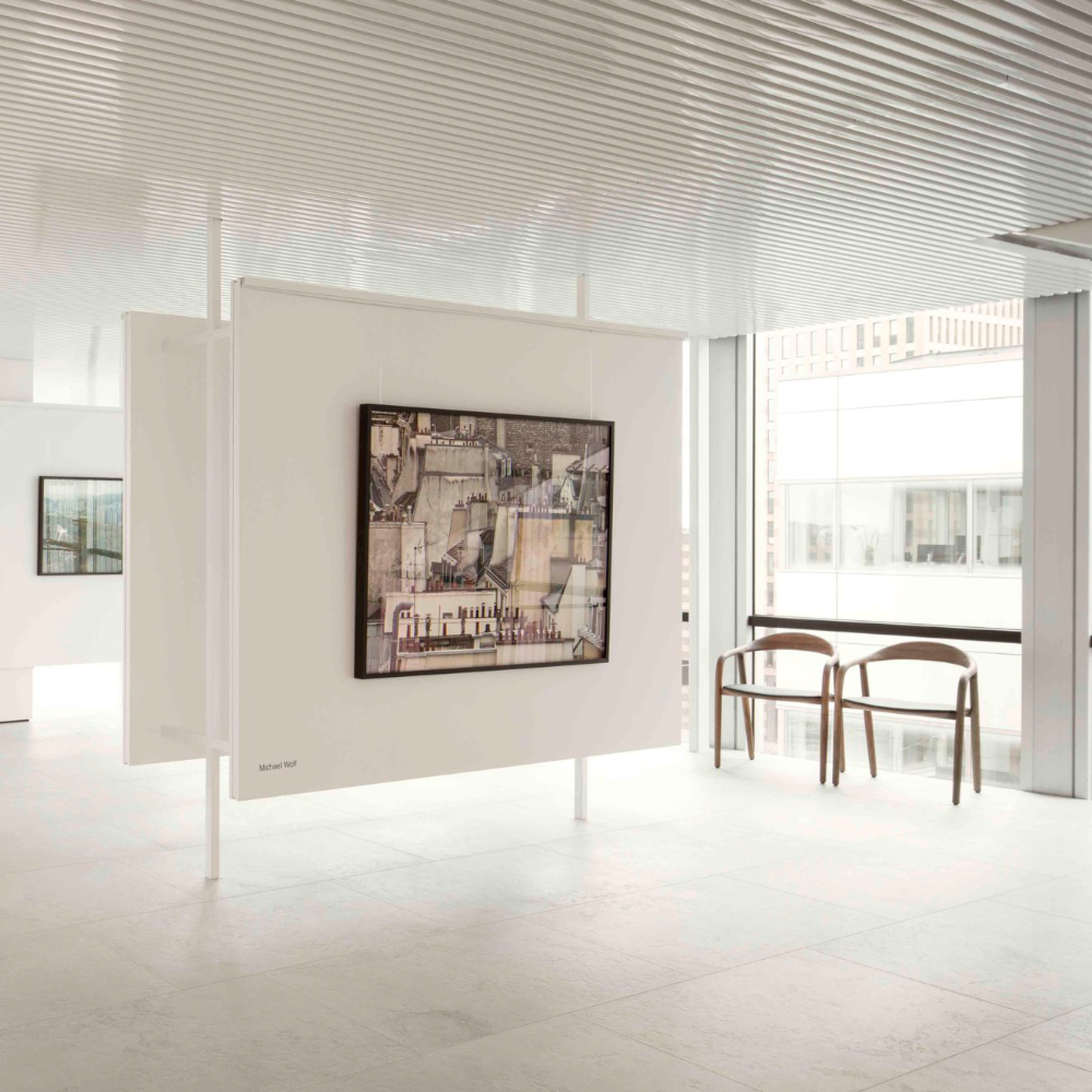 A minimalist art gallery interior showcases a modern painting on a suspended white panel, with natural light streaming through large windows. In the background, two NEVA Easy Chairs by Artisan feature an ergonomic design against the white walls and floor.