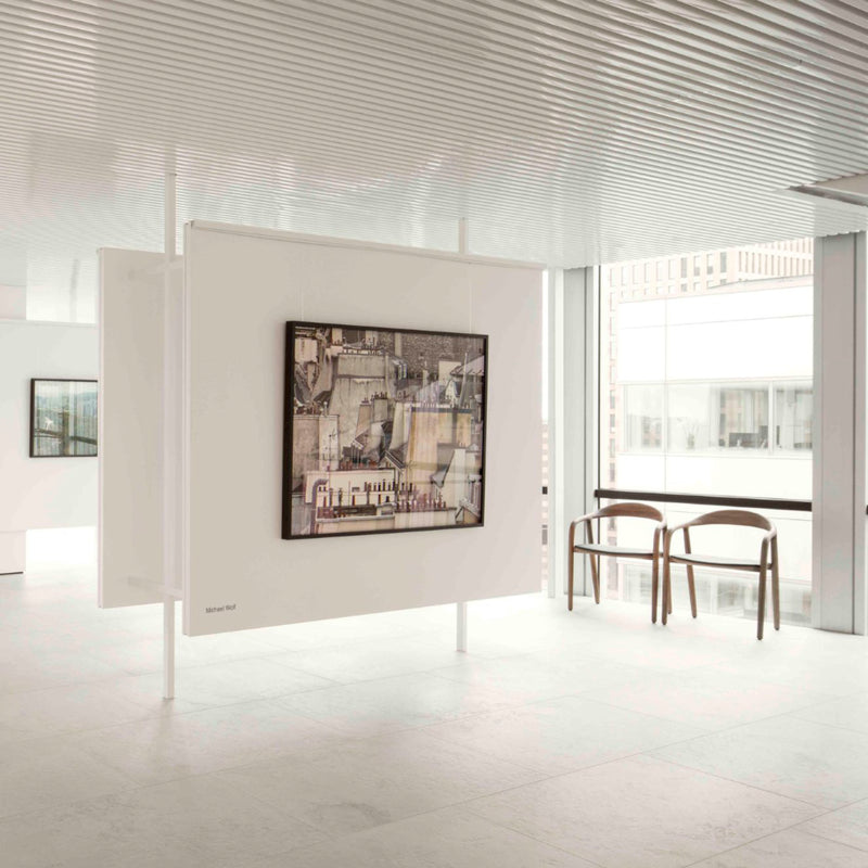 A minimalist art gallery interior showcases a modern painting on a suspended white panel, with natural light streaming through large windows. In the background, two NEVA Easy Chairs by Artisan feature an ergonomic design against the white walls and floor.