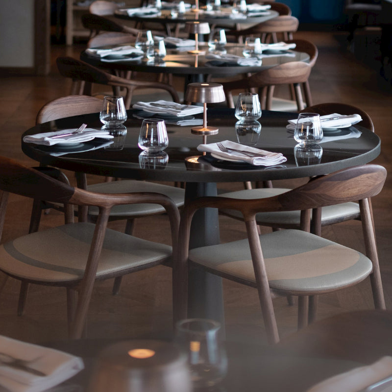 In a minimalist restaurant setting, round wooden tables feature the Artisan NEVA Easy Chair, combining ergonomic design with style. Each table is set with four water glasses, a small lamp, and neatly folded white napkins. Cushioned seats add to the warm and inviting atmosphere.