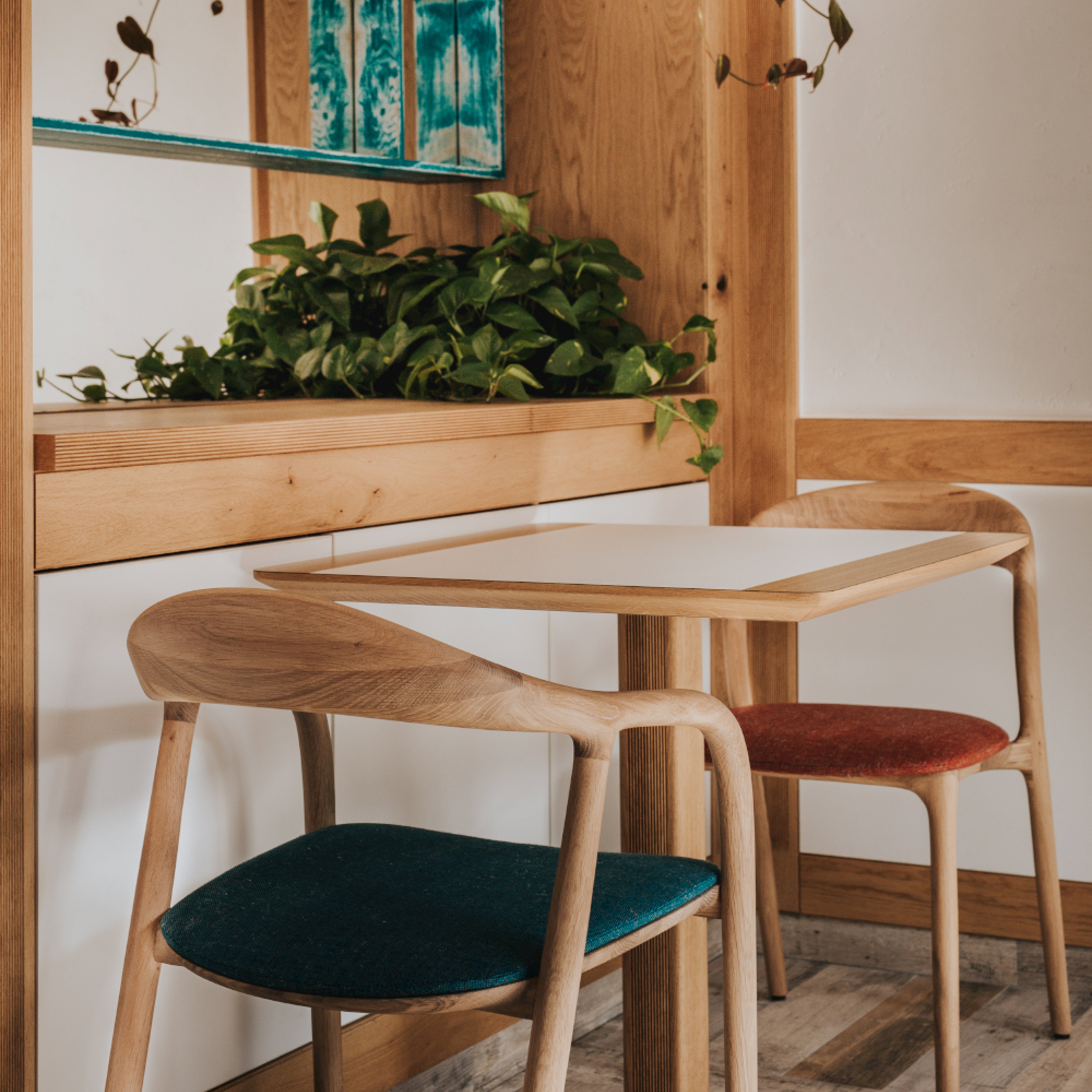A cozy cafe corner features a small wooden table, crafted by Artisan carpentry, with two NEVA light chairs adorned with teal and rust cushions. Wooden shelves hold lush green plants for a refreshing touch. The light wood floor complements the warm tones of this charming collection space.