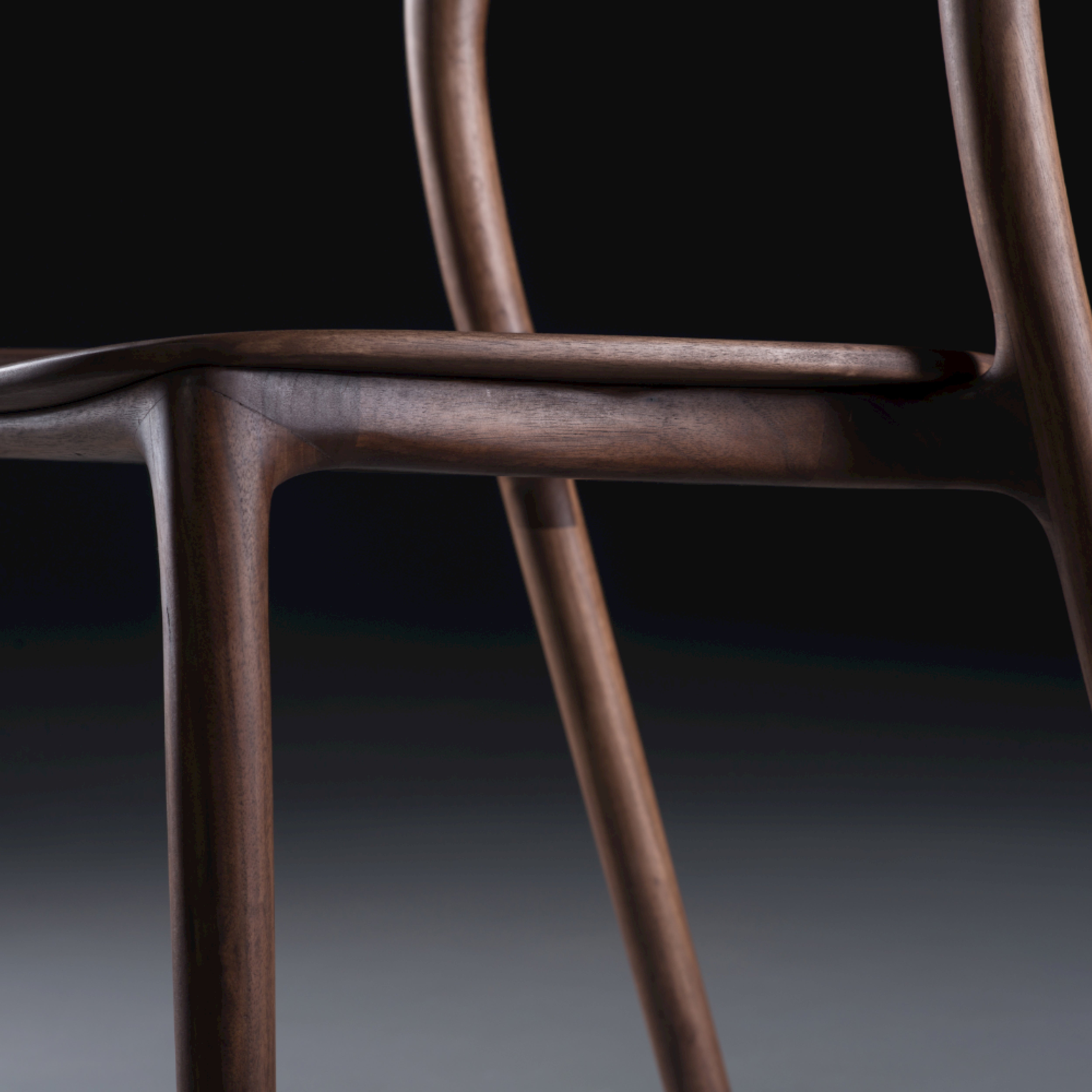 A close-up of the Neva light chair by Artisan features its smooth, curved armrest and backrest with a rich dark finish. The dark background highlights the chairs elegant craftsmanship and sleek artisanal carpentry design.