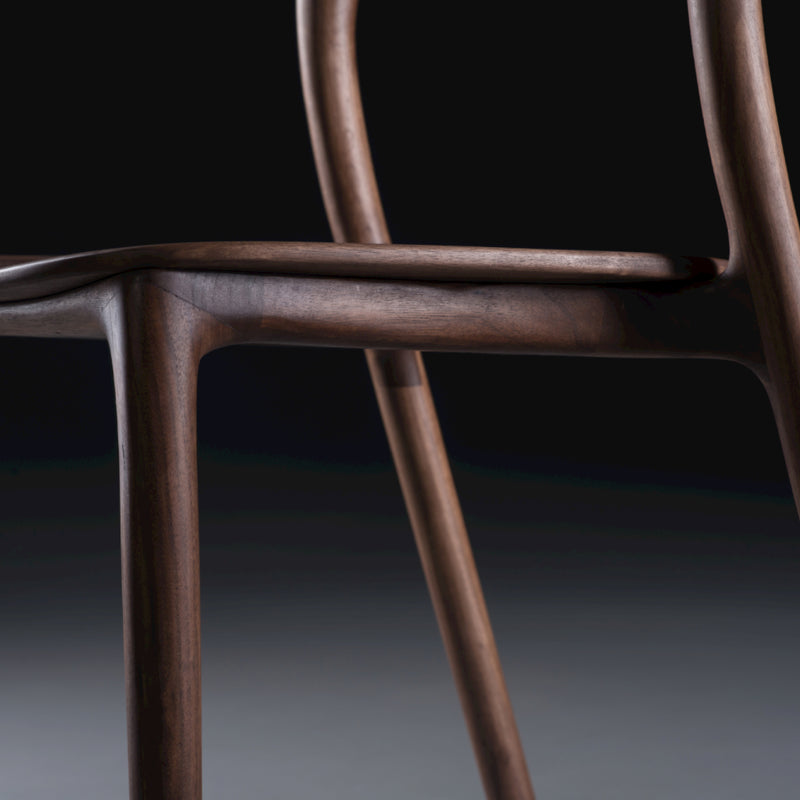 A close-up of the Neva light chair by Artisan features its smooth, curved armrest and backrest with a rich dark finish. The dark background highlights the chairs elegant craftsmanship and sleek artisanal carpentry design.