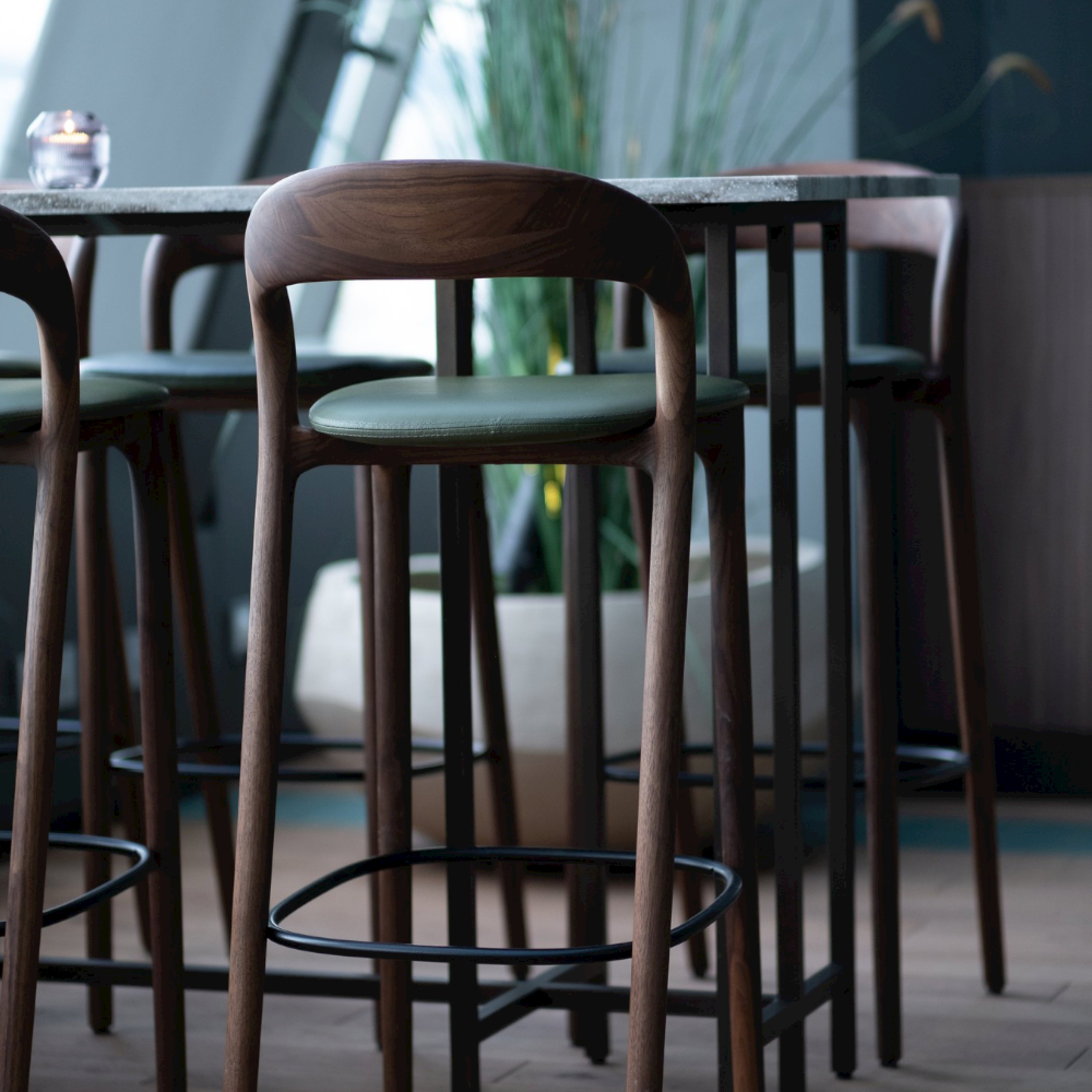 Artisans NEVA light barstools, featuring wooden backs and green cushioned seats, are arranged around a high table in a stylish interior. Indoor plants and soft lighting enhance the chic, cozy atmosphere, highlighting the blend of artisan carpentry and modern technology.