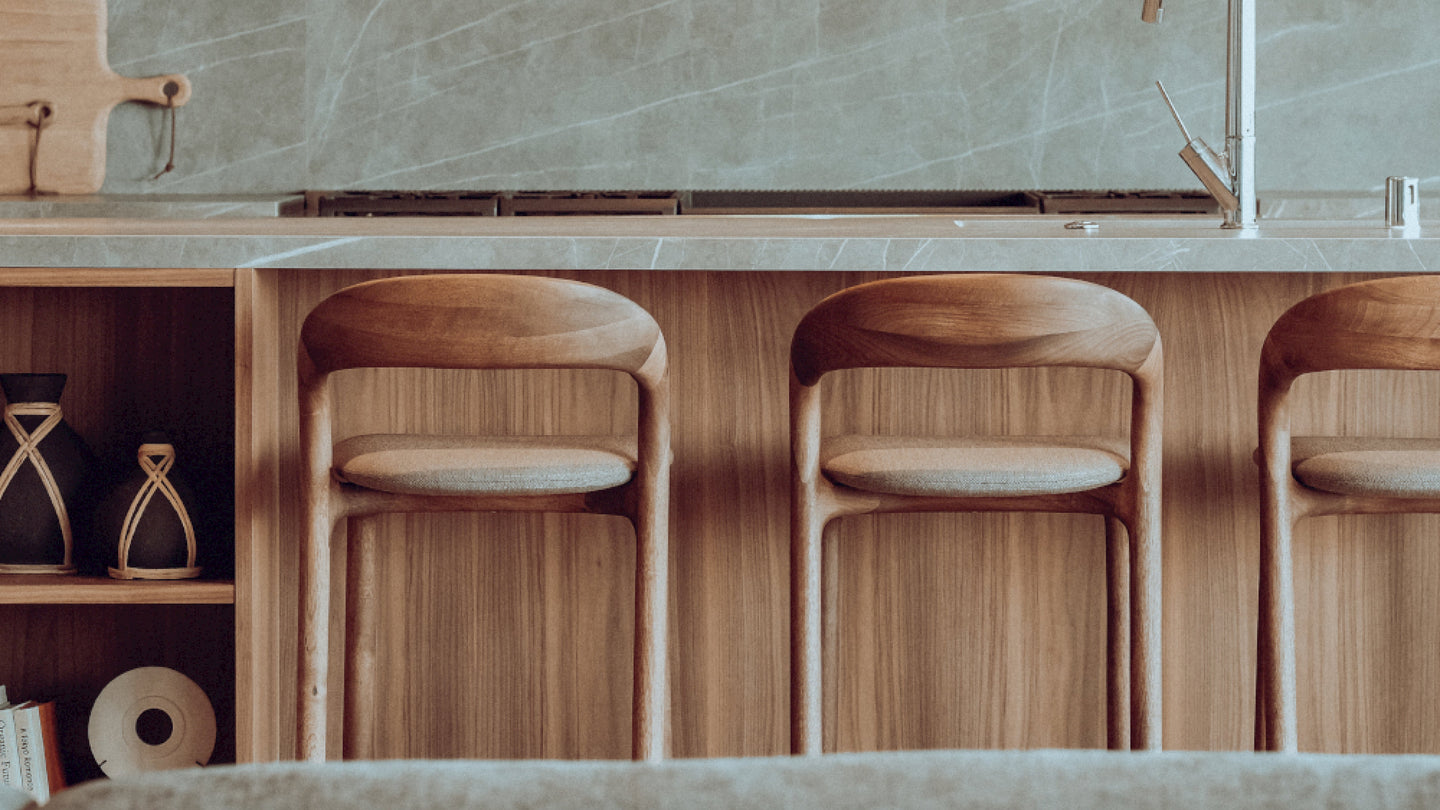 A modern kitchen showcases Artisan craftsmanship with wooden cabinets and a marble backsplash. Three NEVA light barstools align at a counter featuring a sleek metal faucet, embodying the collections style. A decorative shelf displays books and hourglass ornaments.