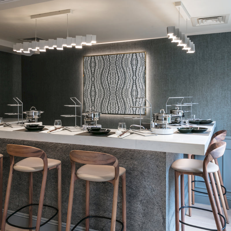 A modern kitchen boasts a large marble island, Artisans NEVA light barstools, sleek lighting fixtures, textured gray wallpaper, patterned artwork, and stylish table settings from the Neva collection on the island counter.