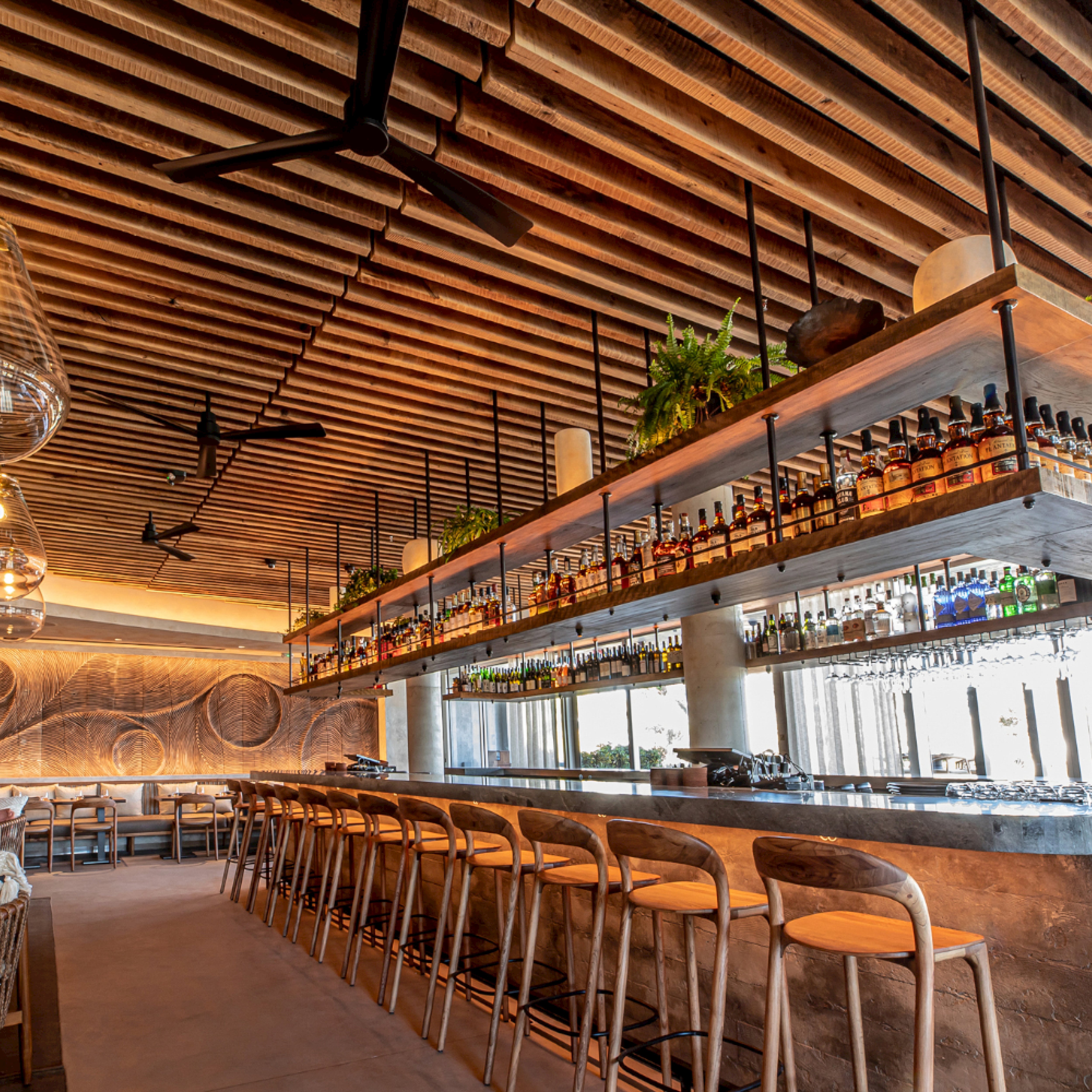 Stylish bar interior featuring the Artisan NEVA light barstool, complemented by a wooden ceiling and floor, a long counter with wooden stools, bottle-stocked shelves, and plants. Warm lighting enhances the cozy atmosphere with modern touches like ceiling fans and hanging lights.