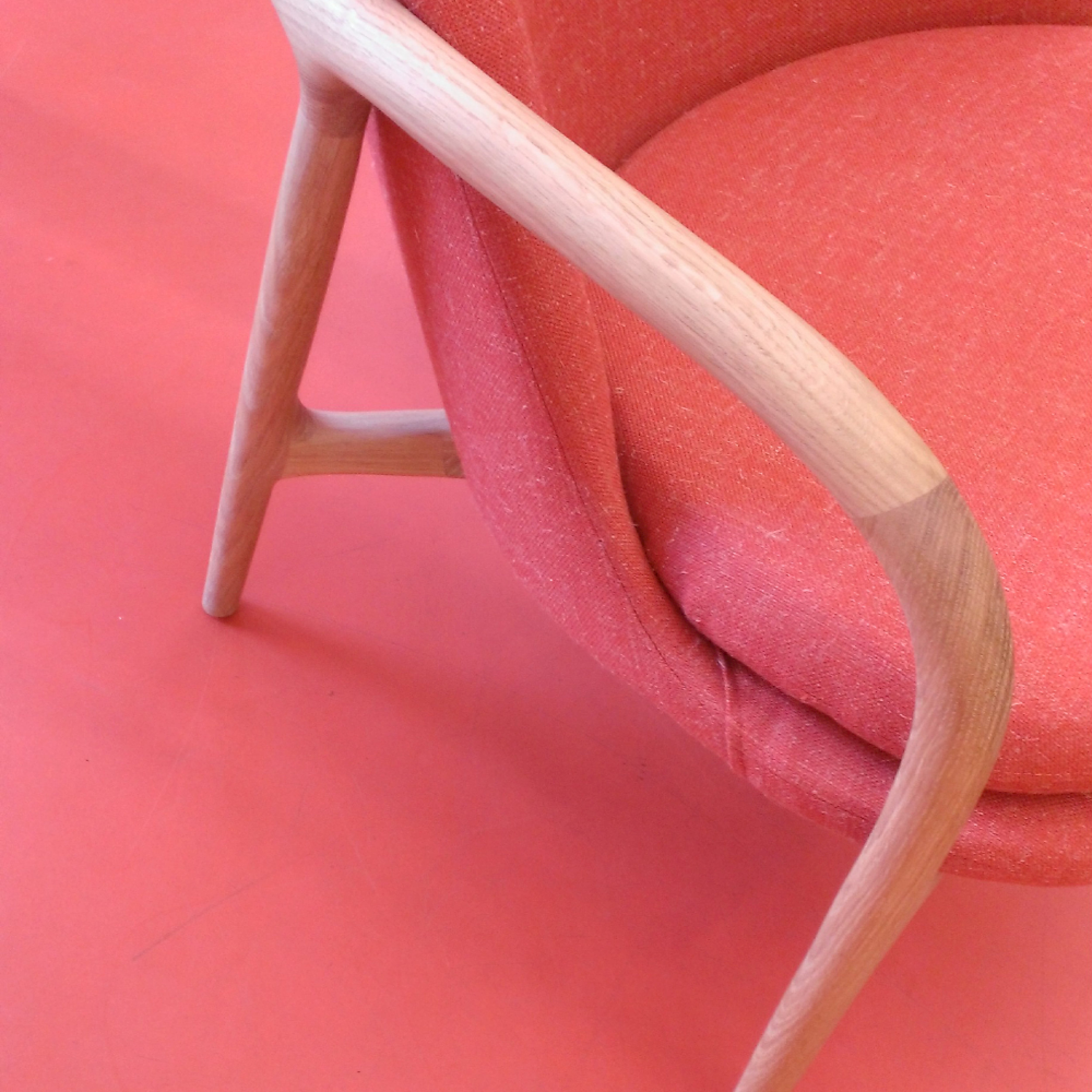 The NEVA Lounge Chair Trimmed by Artisan showcases mid-century modern design with a curved wooden frame and armrests. Its textured red upholstery reflects Scandinavian craftsmanship, pairing seamlessly with a matching red floor.