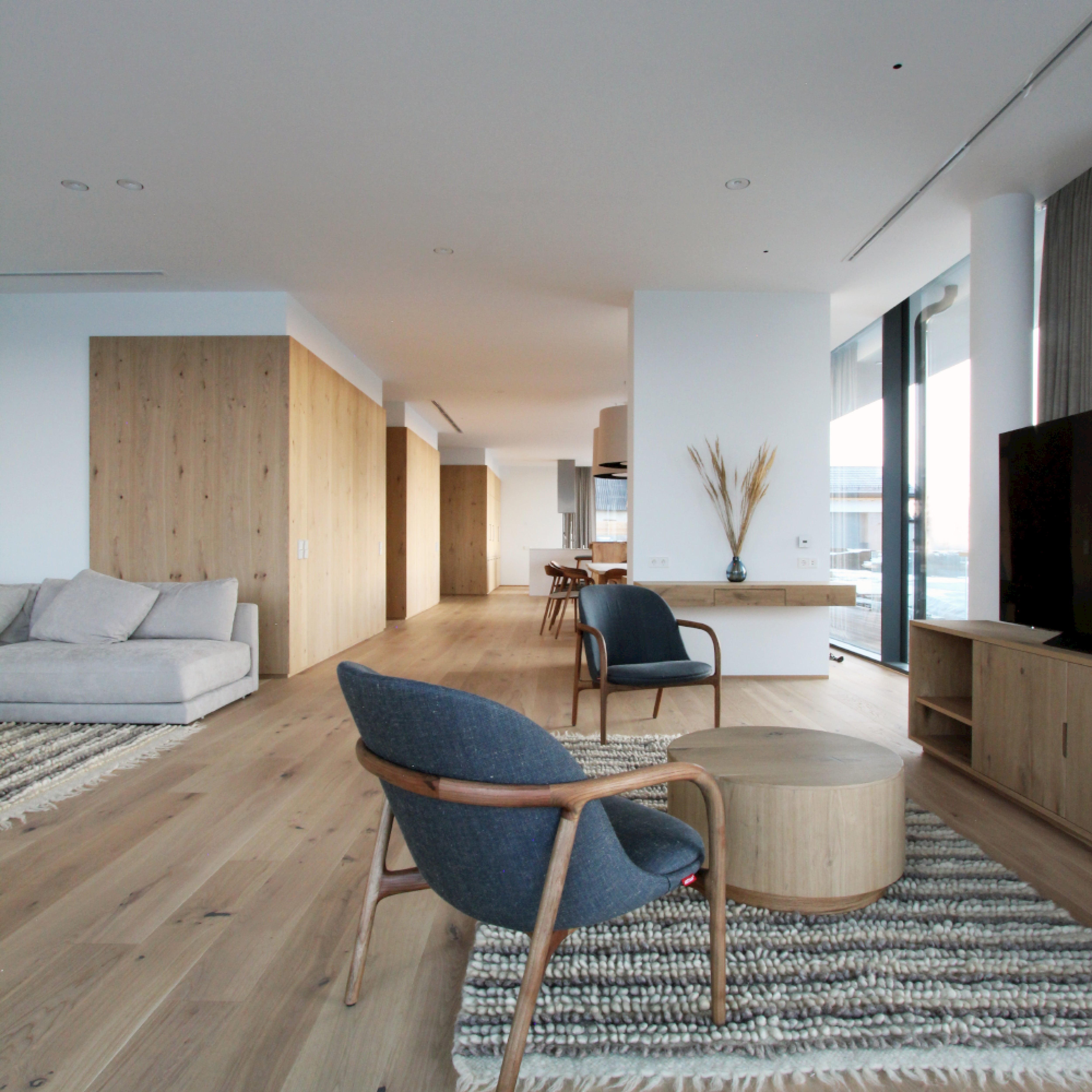 A spacious modern living room features Scandinavian artisanal décor with wooden floors, a gray sofa, and a round wooden coffee table. Two Artisan NEVA Lounge Chairs Trimmed face a large flat-screen TV on a wooden stand. Floor-to-ceiling windows provide a cityscape view in soothing neutral tones.