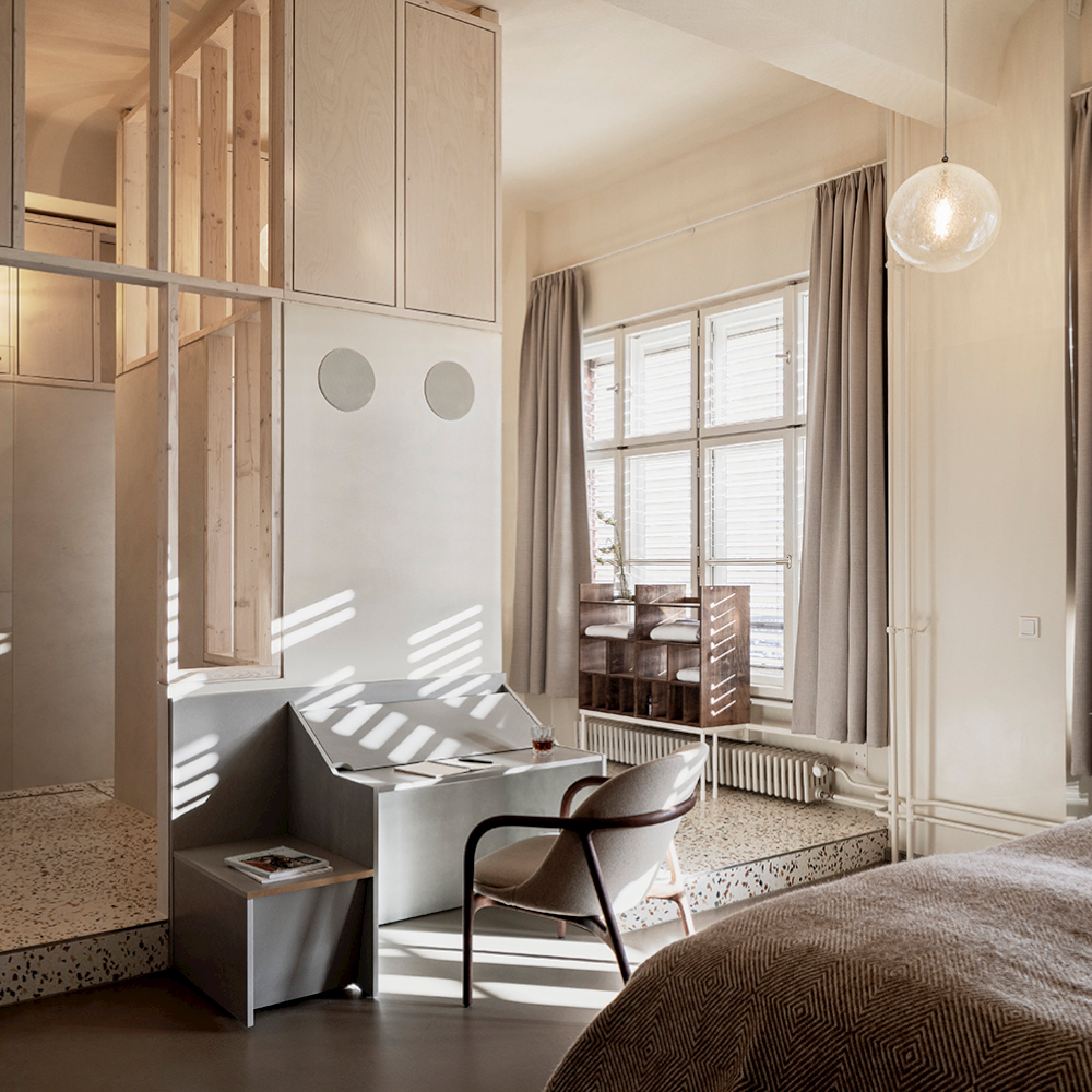 A minimalist, sunlit room with Scandinavian artisanal touches and grey tones includes a textured bed, the modern NEVA Lounge Chair Trimmed by Artisan, a small table with books, large windows with grey curtains, and a unique hanging light fixture.