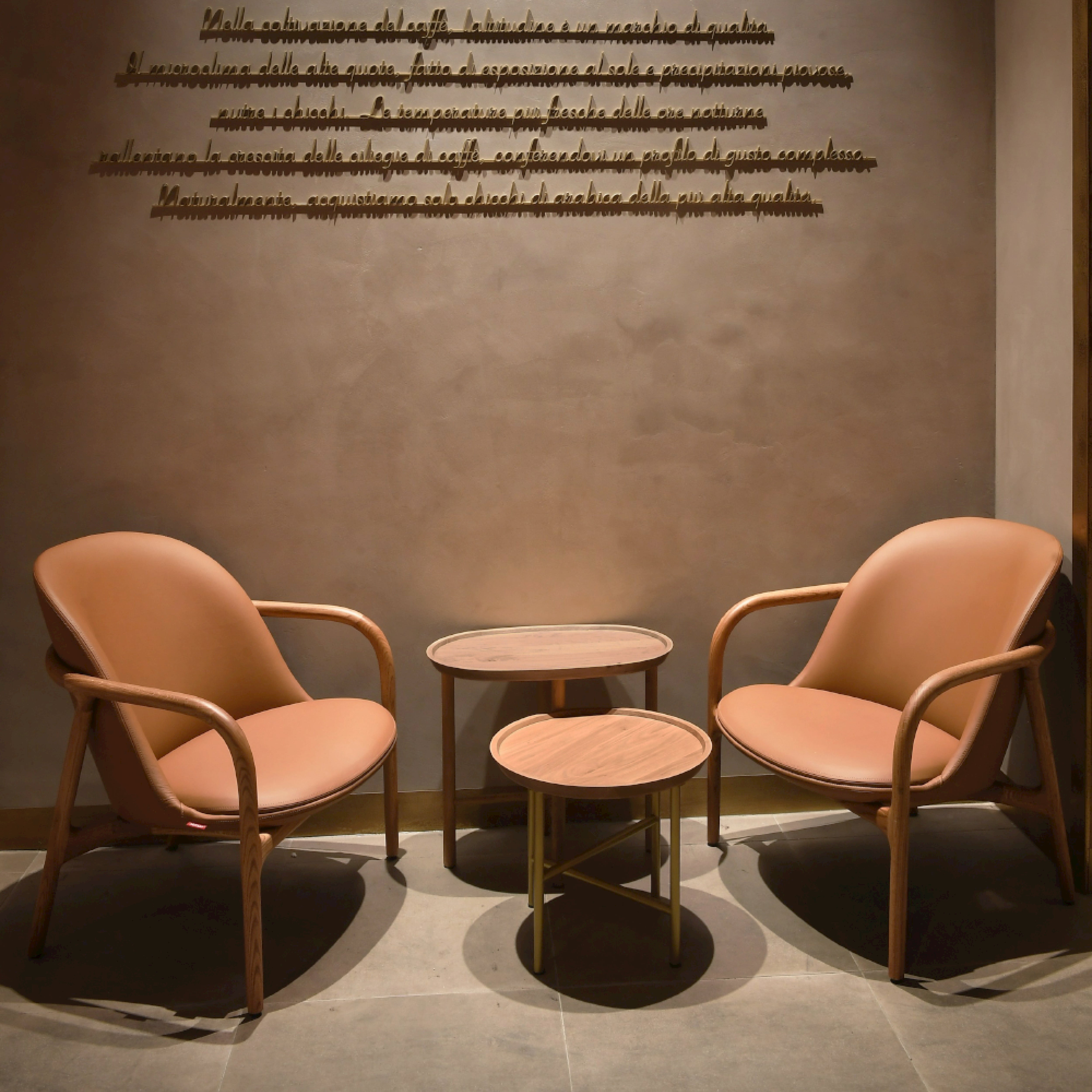 Two tan NEVA Lounge Chairs Trimmed by Artisan, with wooden frames, face each other, separated by nesting round wooden tables of varying heights. The taupe wall behind displays black cursive script, creating a Scandinavian artisanal vibe with a muted modern aesthetic.