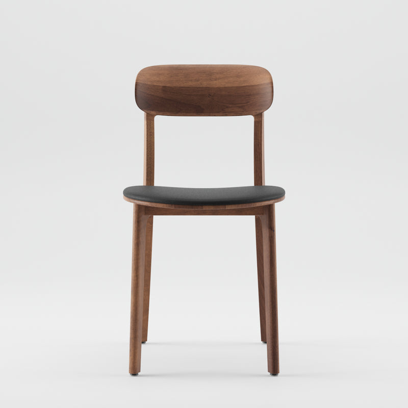 The Artisan TANKA Chair Upholstered showcases a minimalist design with a curved solid wood backrest and a black cushioned seat. Its lightweight wooden frame with four straight legs is elegantly presented against a plain white backdrop.