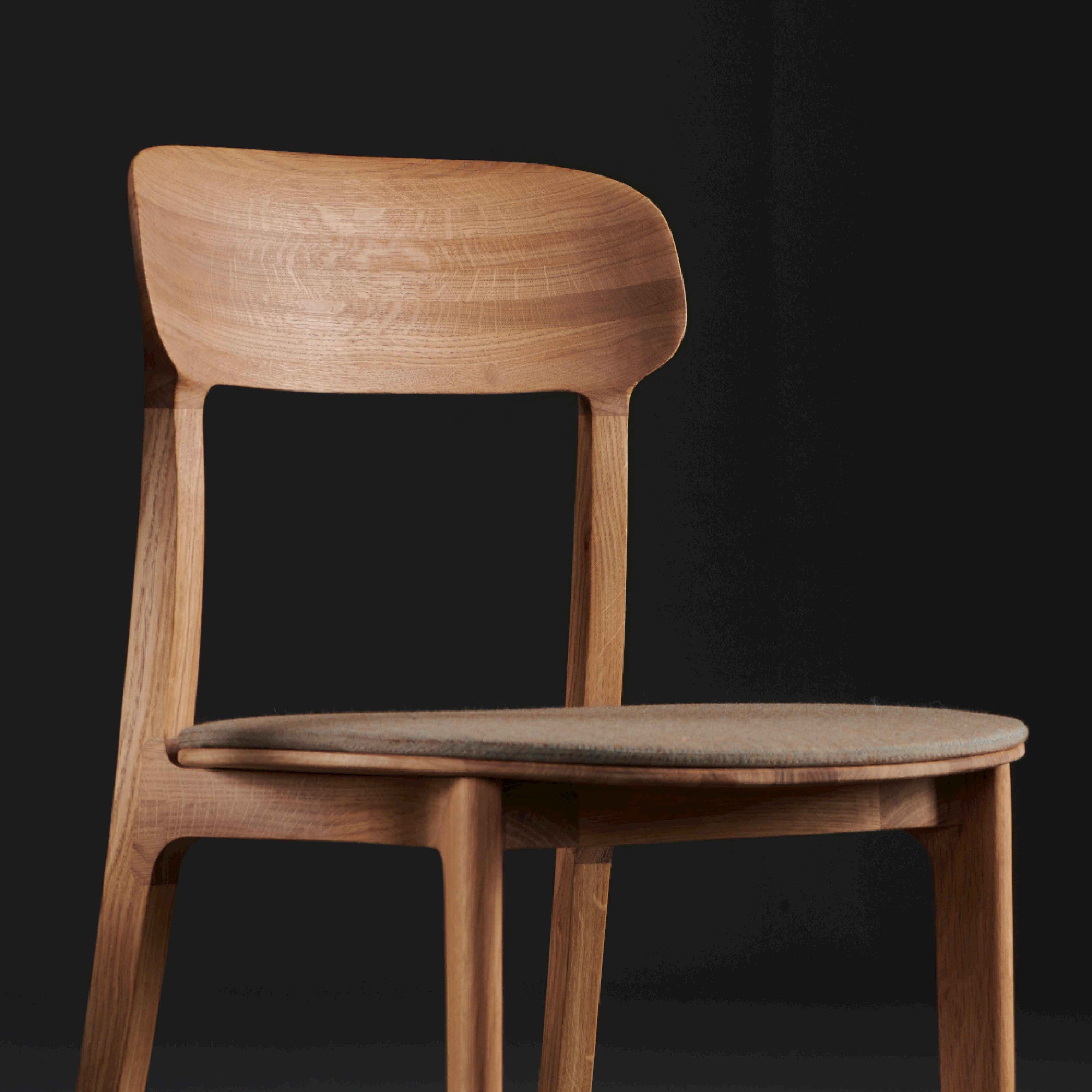 Against a dark background, an Artisan TANKA Chair is positioned, featuring a curved backrest and cushioned seat. Its smooth, light wood finish and simple yet elegant design emphasize its natural finishes.