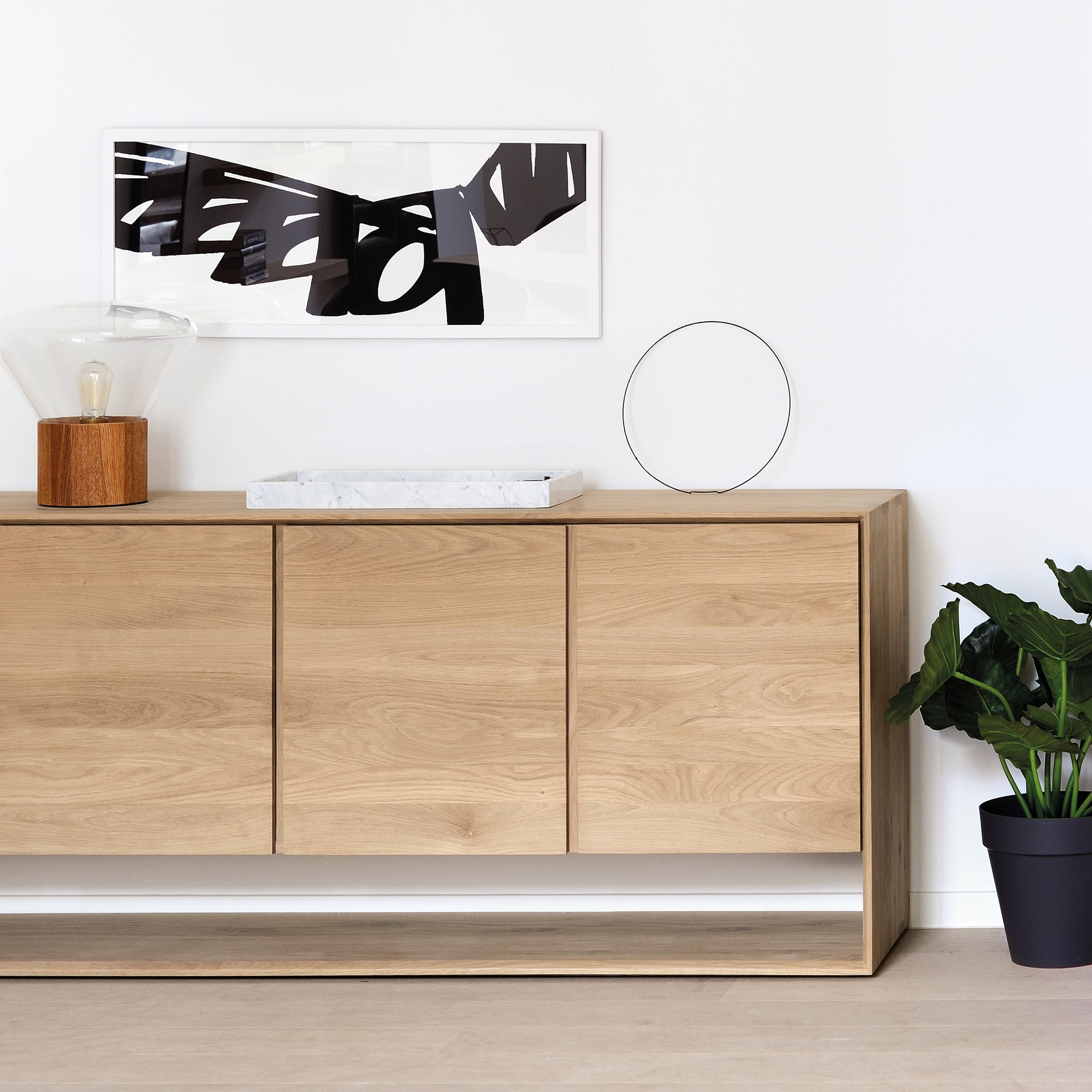 The Ethnicraft Nordic Sideboard stands against a white wall, topped with a lamp, marble tray, and metal ring sculpture. A potted plant is nearby, with abstract black-and-white art displayed above.
