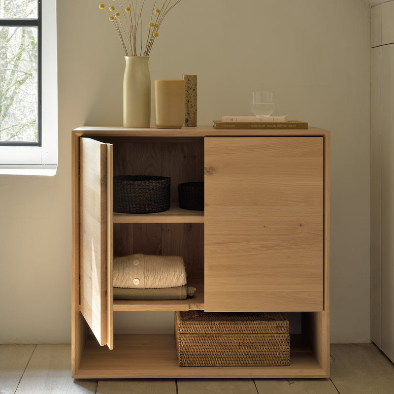 The Nordic Sideboard by Ethnicraft, crafted from light wood, features open shelving with woven baskets and folded towels. On top sit a vase of dried flowers, a book, a small jar, and a glass, all illuminated by sunlight from a nearby window.