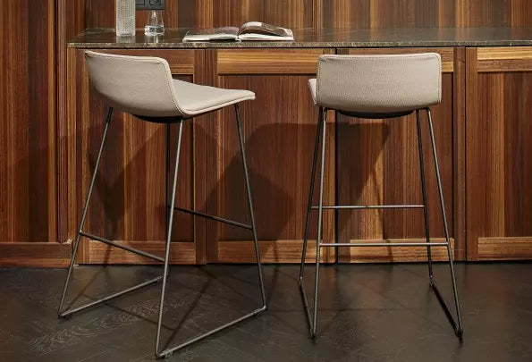 Two Resol Skin Barstool Upholstered chairs, featuring gray cushions and metal legs, are placed at a wooden bar with an open book and glass bottle on top. The setting is framed by wood paneling and dark flooring, ideal for indoor use.