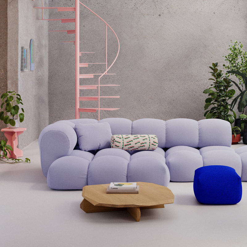 A modern living room features a light purple tufted sectional with a patterned pillow and a blue ottoman. The Objekte unserer Tage Fels Coffee Table, noted for its craftsmanship, is central amid nearby plants. A pink spiral staircase ascends the textured gray wall, adding sculptural beauty.