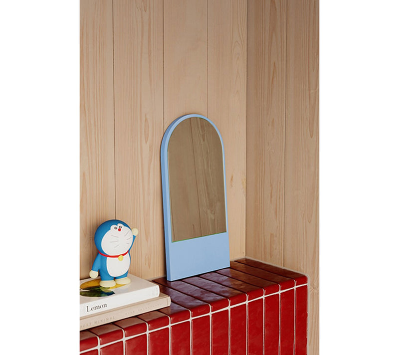 A small blue Objekte unserer Tage FRIEDRICH Mini toy figure stands on a stack of books beside a light blue arched Friedrich Mini Mirror, set against a red-tiled ledge and wooden wall panels adorned with subtle floral patterns.
