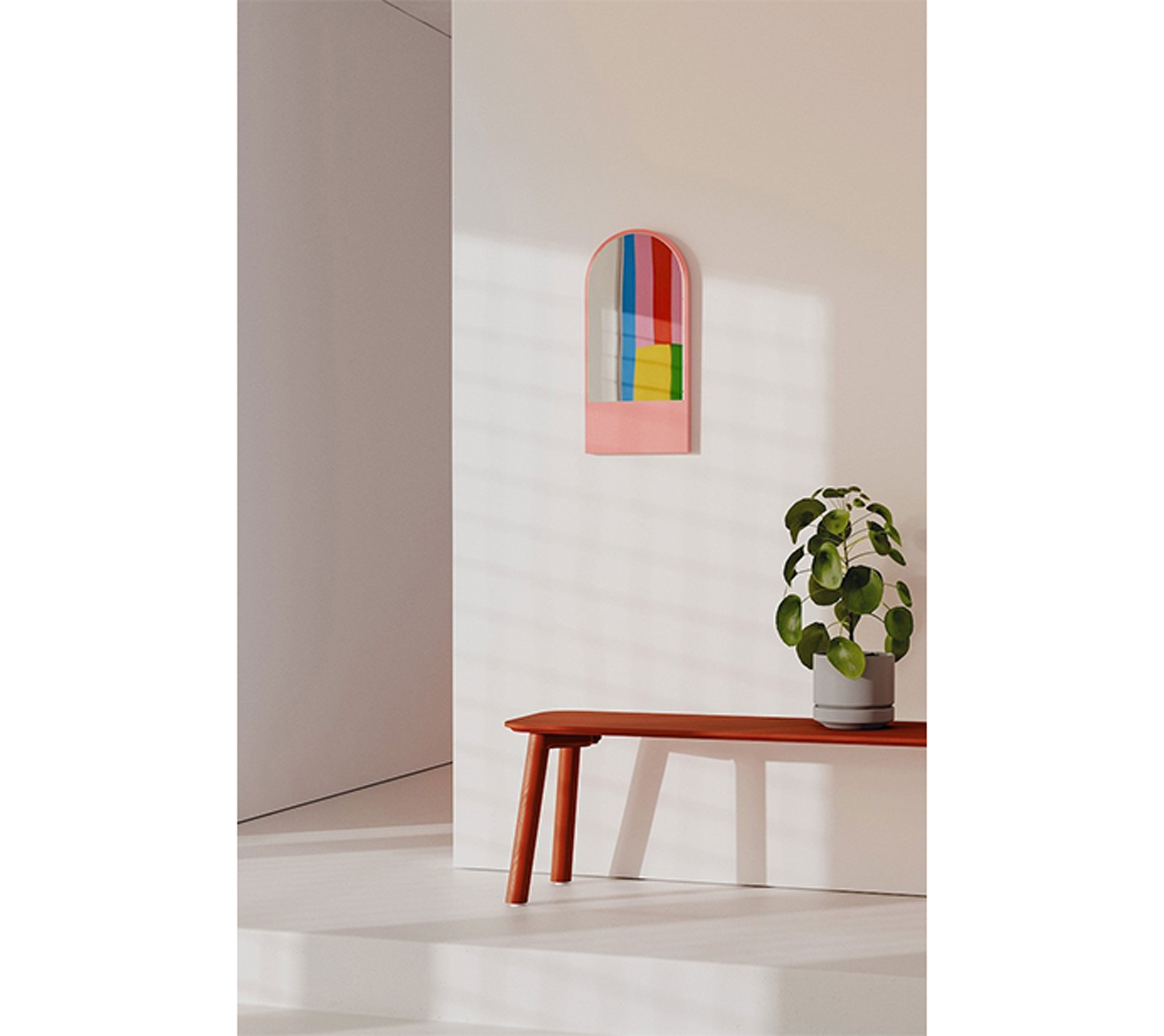 A minimalist room with a white wall showcases the Friedrich Mini Mirror by Objekte unserer Tage, featuring expressive colors in an abstract arch design. Below, a potted plant rests on a sleek red bench with floral wood grain. Soft light casts gentle shadows, creating serenity.