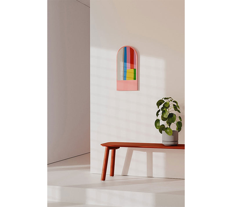 A minimalist room with a white wall showcases the Friedrich Mini Mirror by Objekte unserer Tage, featuring expressive colors in an abstract arch design. Below, a potted plant rests on a sleek red bench with floral wood grain. Soft light casts gentle shadows, creating serenity.