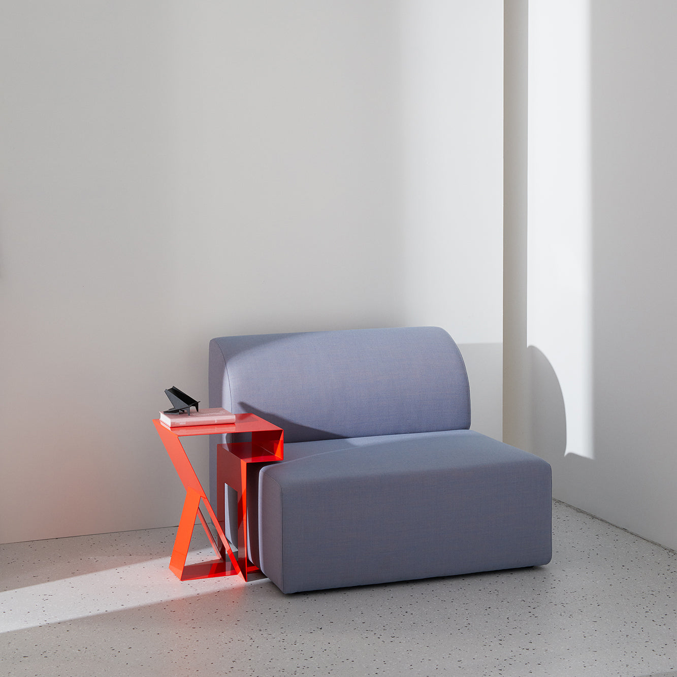 A minimalist room features the Objekte unserer Tages Weber Modular Sofa in light gray, illustrating sustainable awareness, next to a small, angular translucent red side table holding a smartphone and two books. Soft light casts shadows on plain walls and a speckled floor.