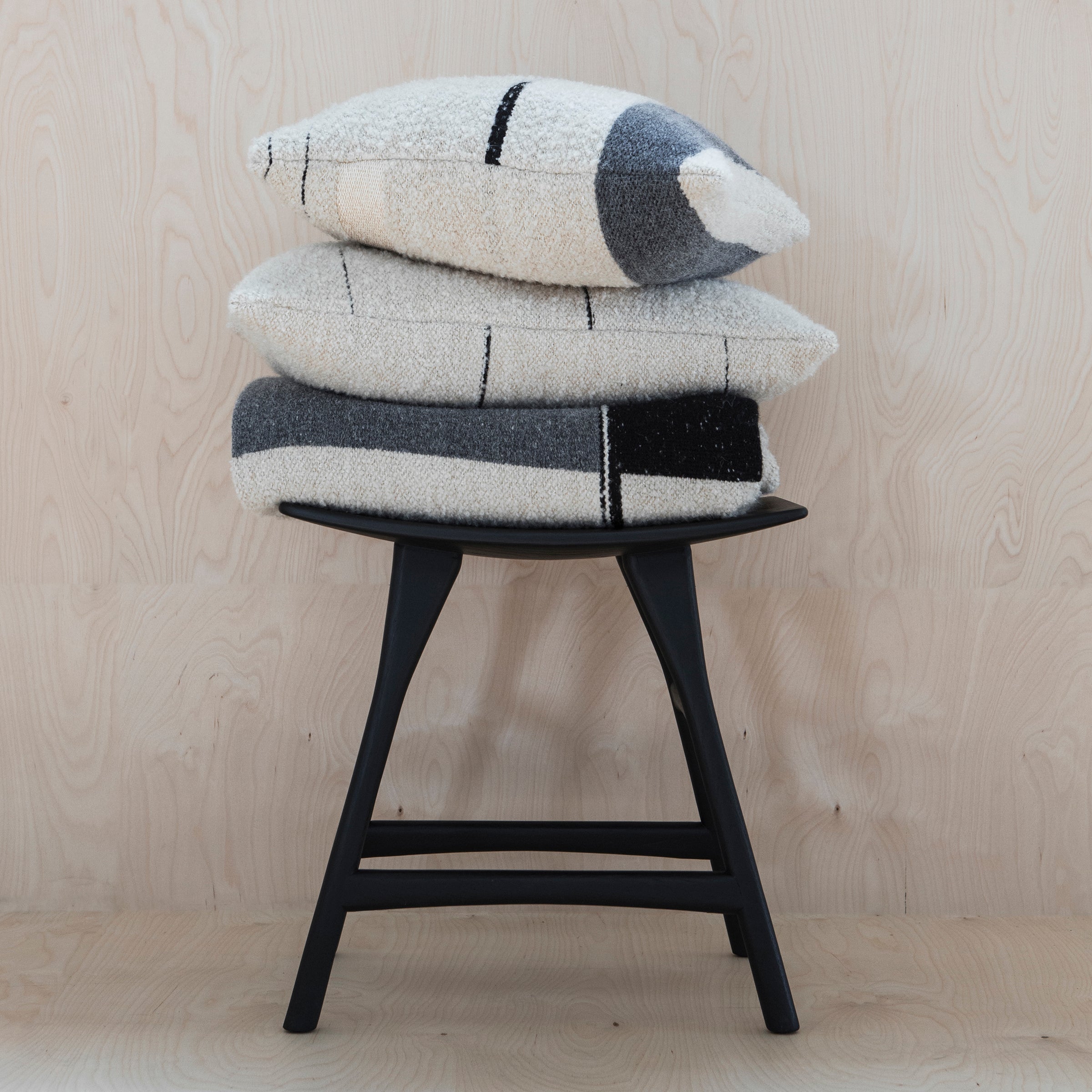 Three textured, cream pillows with black and gray geometric patterns are stacked on the Ethnicraft Osso Stool, crafted from oak wood and set against a light wood background.