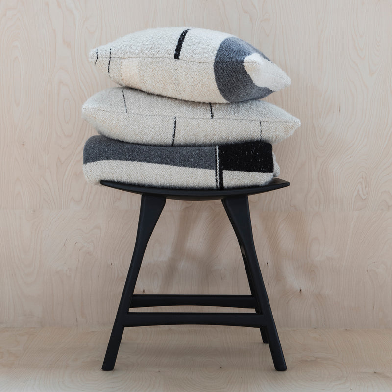 Three textured, cream pillows with black and gray geometric patterns are stacked on the Ethnicraft Osso Stool, crafted from oak wood and set against a light wood background.